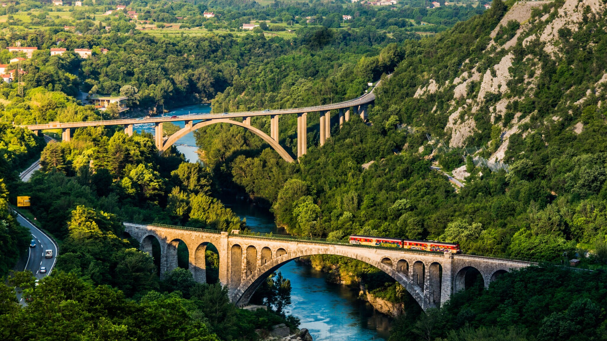 Regionalzug fährt über einen steinernen Viadukt über einen Fluss in einer grünen, bergigen Landschaft in Slowenien.