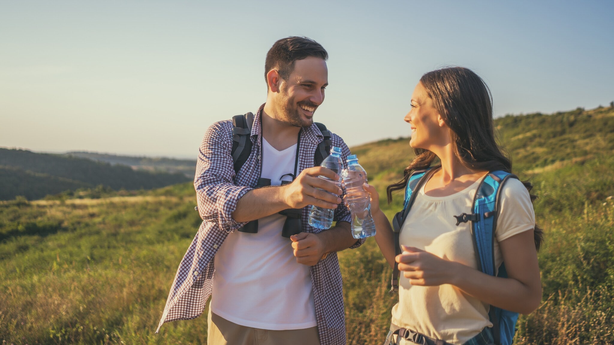 Zwei Personen stehen auf einer Wiese, der Mann reicht der Frau eine Wasserflasche. Beide tragen Rucksäcke und lächeln. Zwei Personen stehen auf einer Wiese, der Mann reicht der Frau eine Wasserflasche. Beide tragen Rucksäcke und lächeln.
