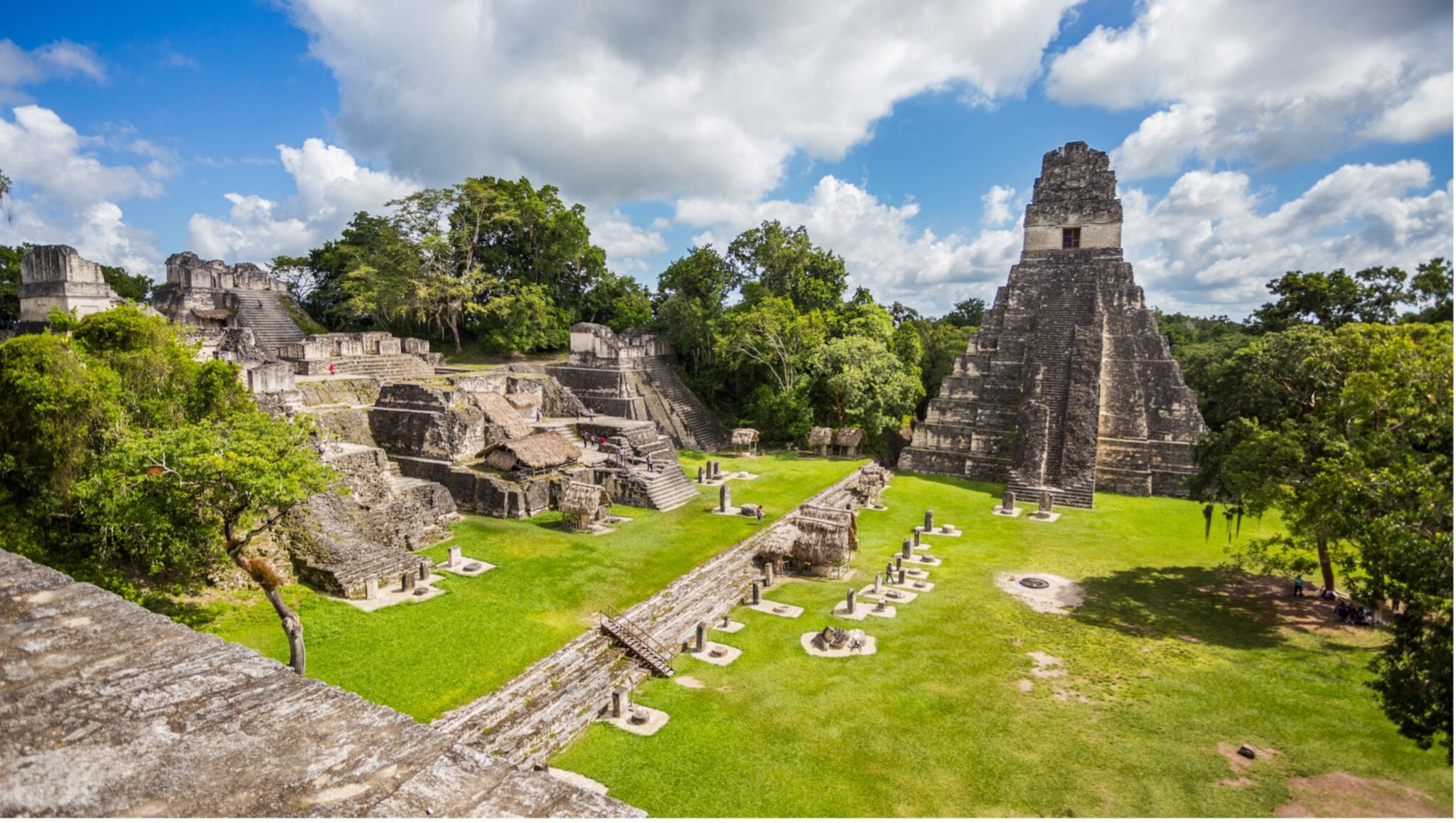 Maya-Tempelanlage mit Stufenpyramide und mehreren Steinstrukturen umgeben von grünem Gras und Bäumen unter blauem Himmel mit Wolken