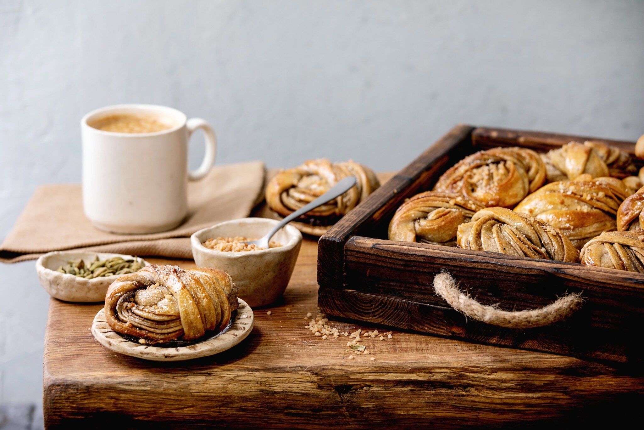 Holztisch mit einem Tablett voll schwedischer Kanelbullar, Keramikschalen mit Kardamom und gehackten Nüssen sowie eine Tasse Kaffee.