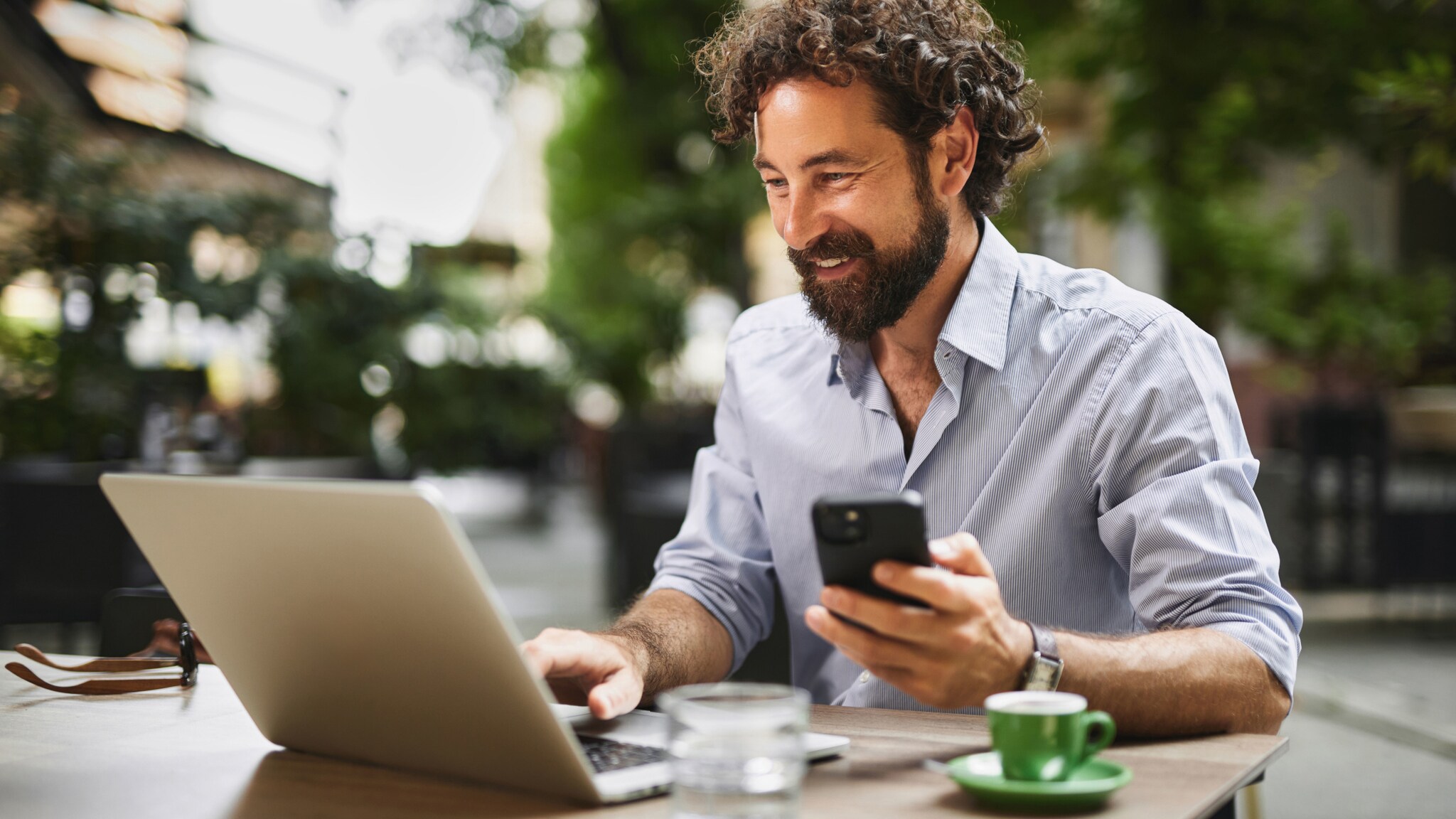 Ein Mann im legeren Hemd arbeitet an einem Laptop, neben sich eine Espressotasse, in einer Hand hält er ein Smartphone.