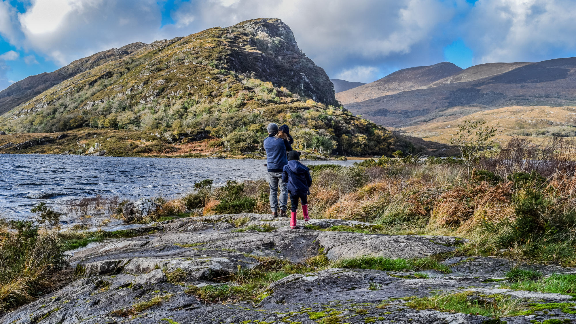 Vater mit Kindern stehen in Outdoor-Kleidung an einem Seeufer mit felsigem Untergrund, im Hintergrund bewachsene Hügel und Berge unter bewölktem Himmel. Vater mit Kindern stehen in Outdoor-Kleidung an einem Seeufer mit felsigem Untergrund, im Hintergrund bewachsene Hügel und Berge unter bewölktem Himmel.