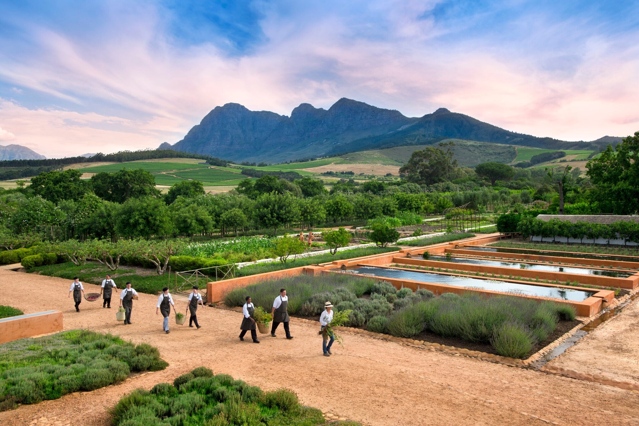 Großes Gartenareal mit verschiedenen Beeten, durch das eine Personengruppe mit der Ernte läuft, im Hintergrund hügelige Landschaft im Morgenlicht Großes Gartenareal mit verschiedenen Beeten, durch das eine Personengruppe mit der Ernte läuft, im Hintergrund hügelige Landschaft im Morgenlicht