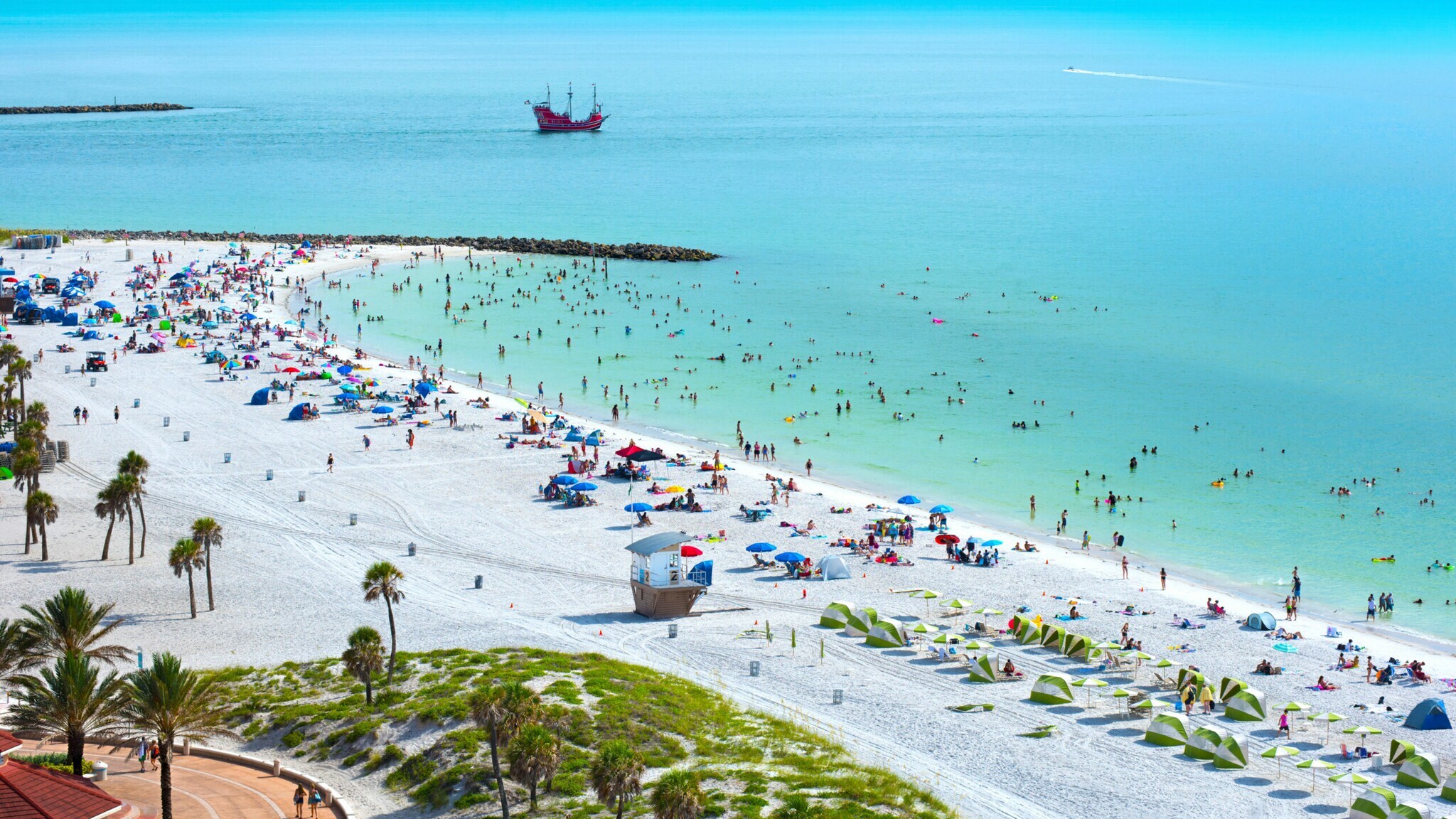 Belebter, breiter Strand mit weißem Sand an türkisblauem Wasser.