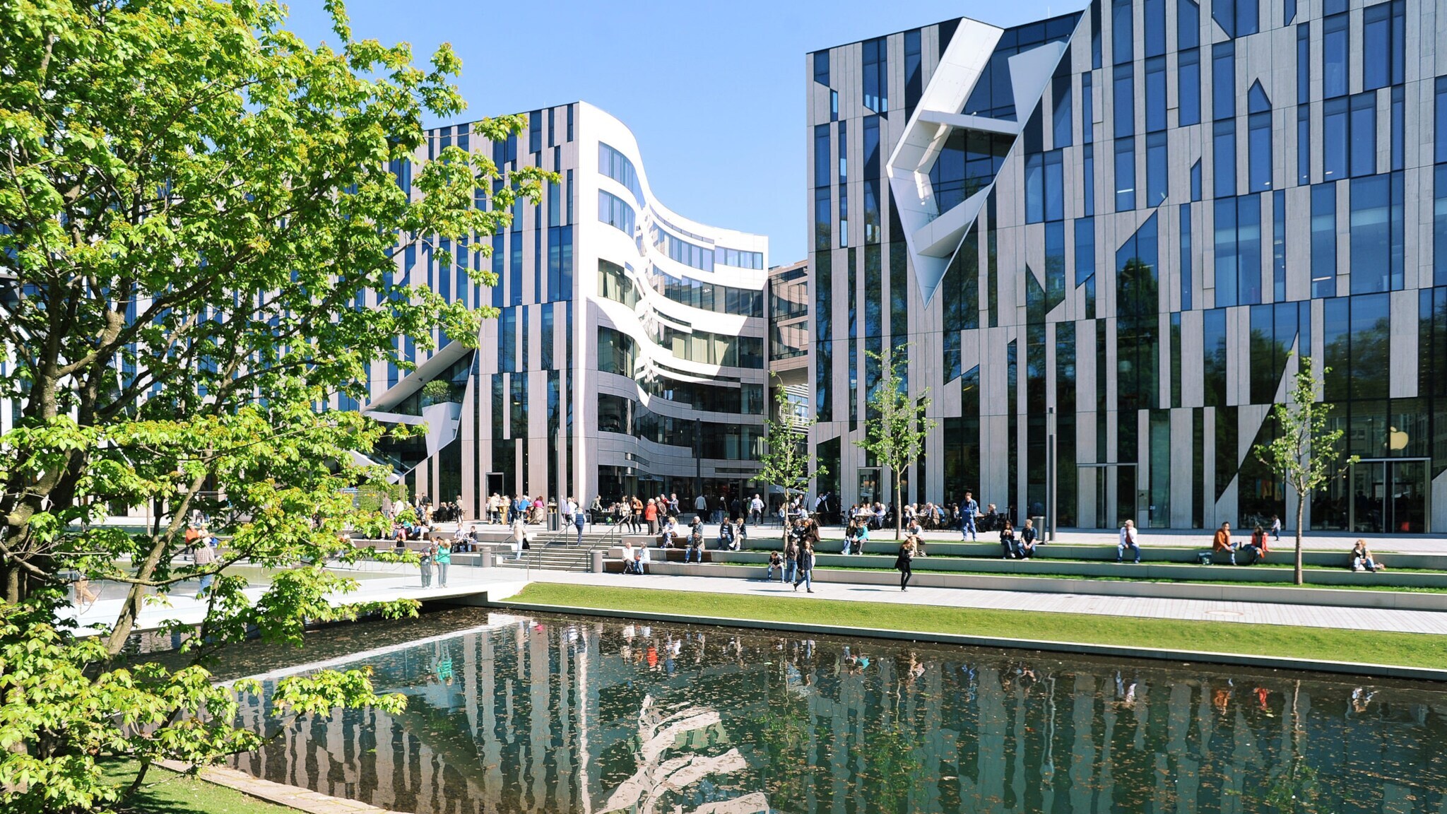 Moderne Bürogebäude mit Glasfassaden am Wasserbecken im Kö-Bogen Düsseldorf, Menschen sitzen und gehen auf der Promenade.
