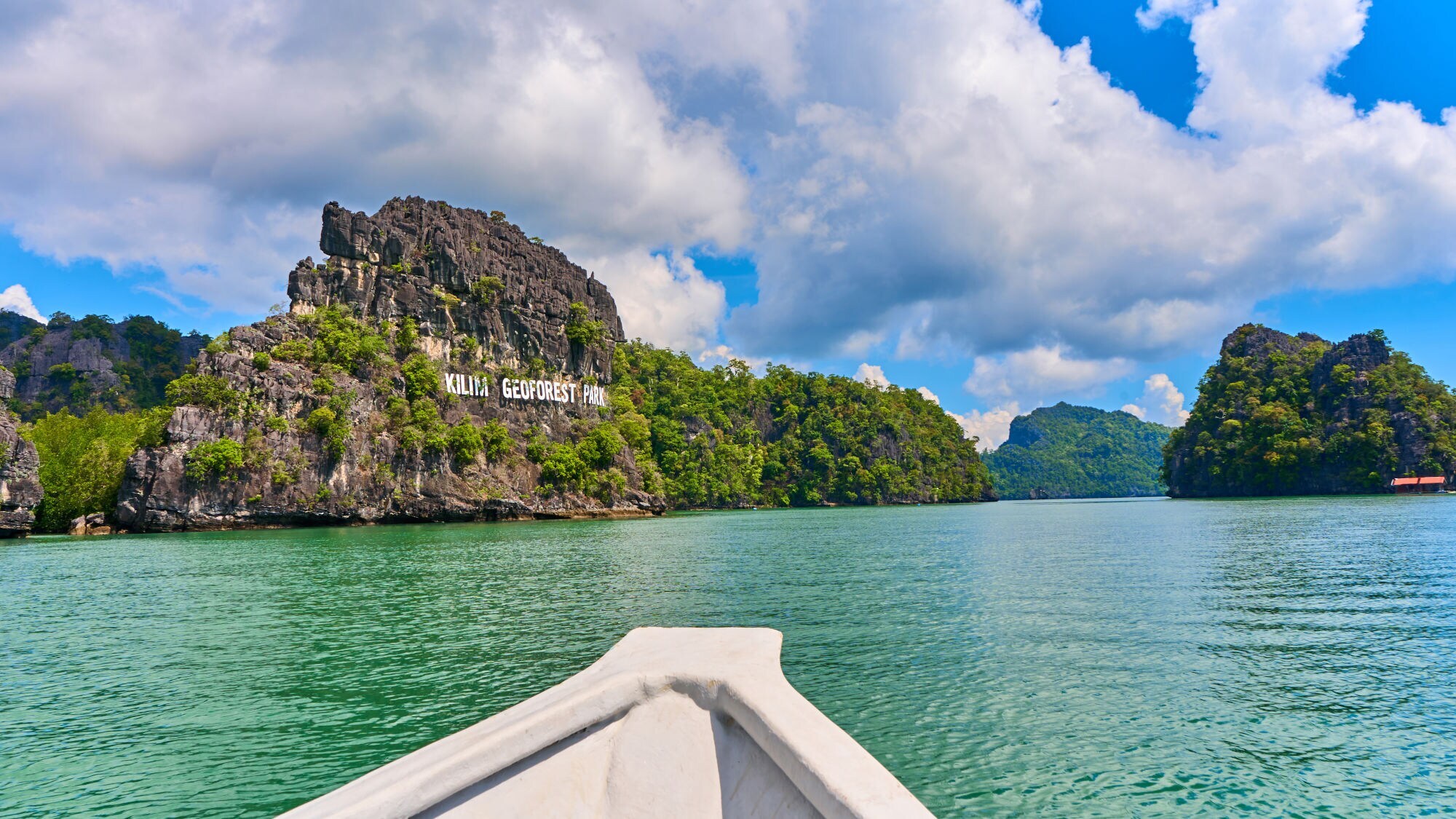 Blick von einem Boot auf grüne Felseninseln mit der Aufschrift 'KILIM GEOPARK' unter blauem Himmel mit Wolken.