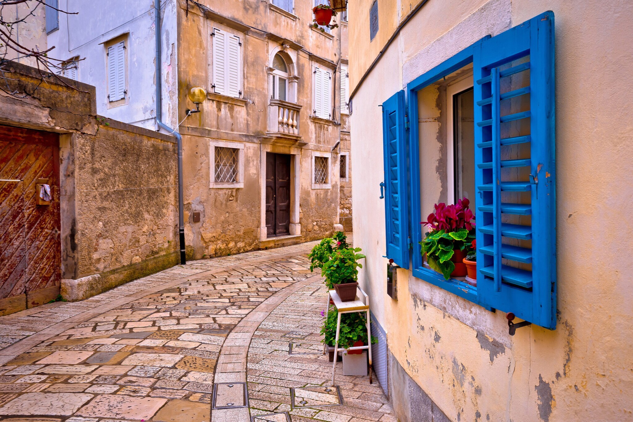 Eine schmale Altstadtgasse, im Vordergrund ein Haus mit blauen Fensterläden und Blumen vor den Fenstern.