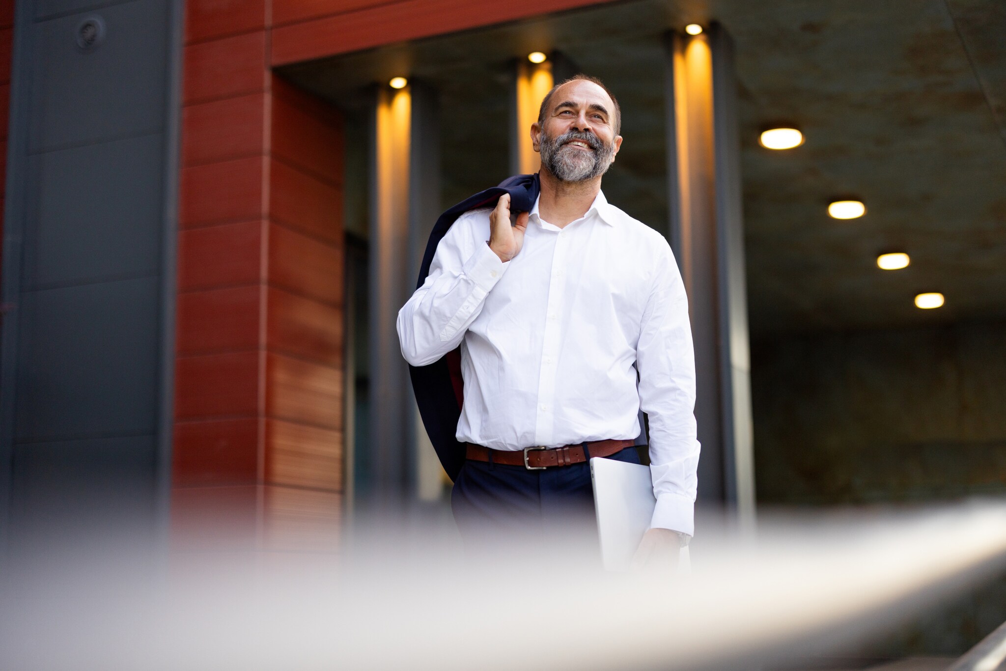 Ein lächelnder Mann im weißen Hemd steht mit einem über die Schulter geworfenen Jackett und Laptop in der Hand vor einem Gebäude.