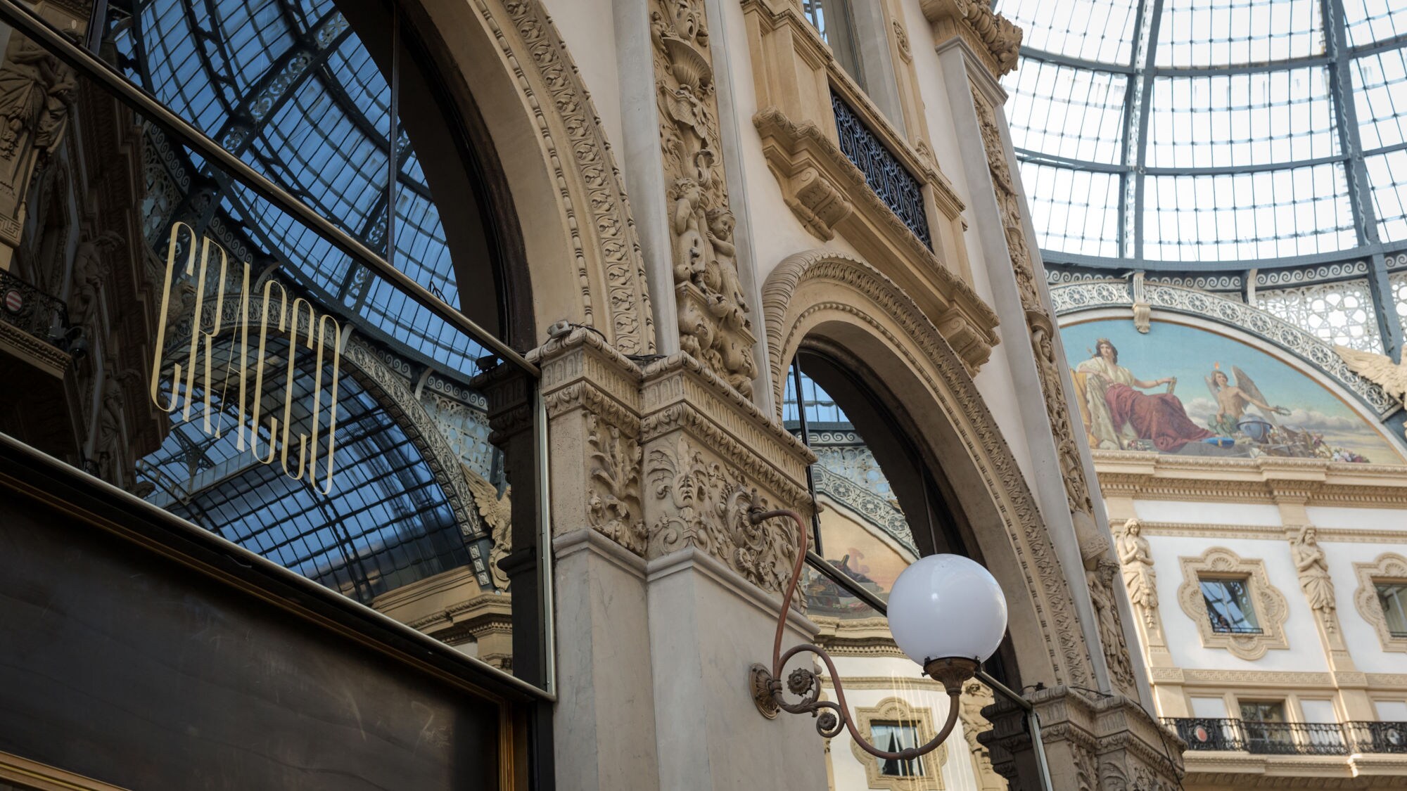 Architekturansicht eines eleganten Gebäudes mit großen Bögen und kunstvollen Verzierungen. Reflektierende Fenster zeigen den Innenraum und eine Wandmalerei.