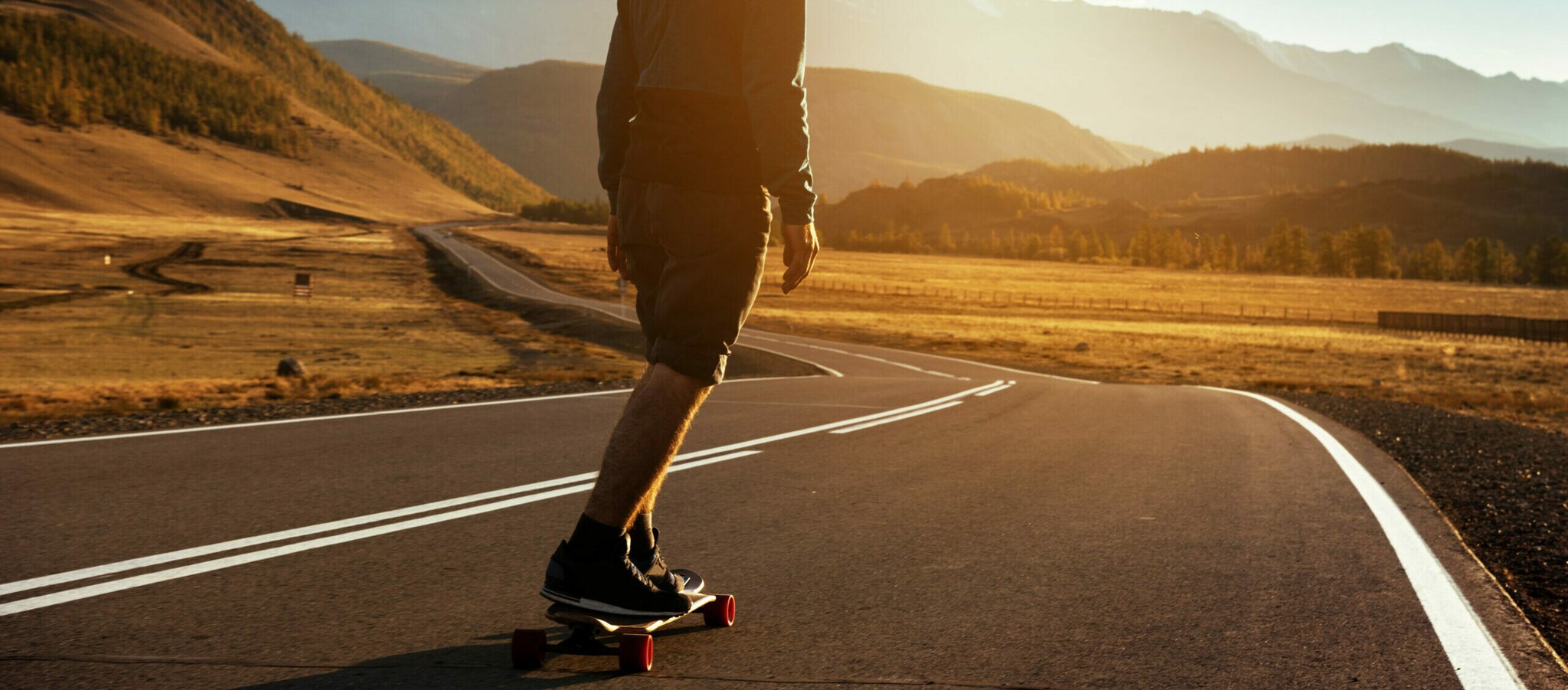 Karge Landschaft vor einer Bergkette, ein Mann fährt auf einem Longboard eine lange, leere Straße entlang.