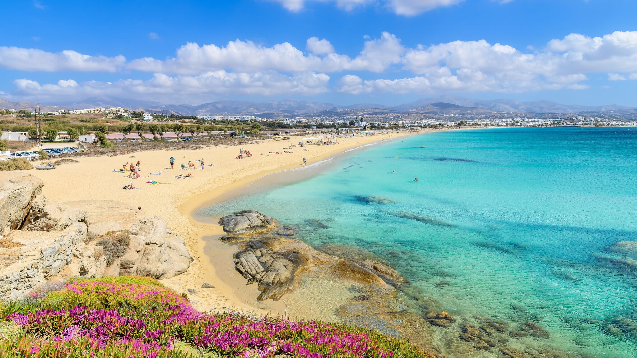 Personen auf einem hellen Sandstrand am türkisblauen Meer in mediterraner Umgebung.