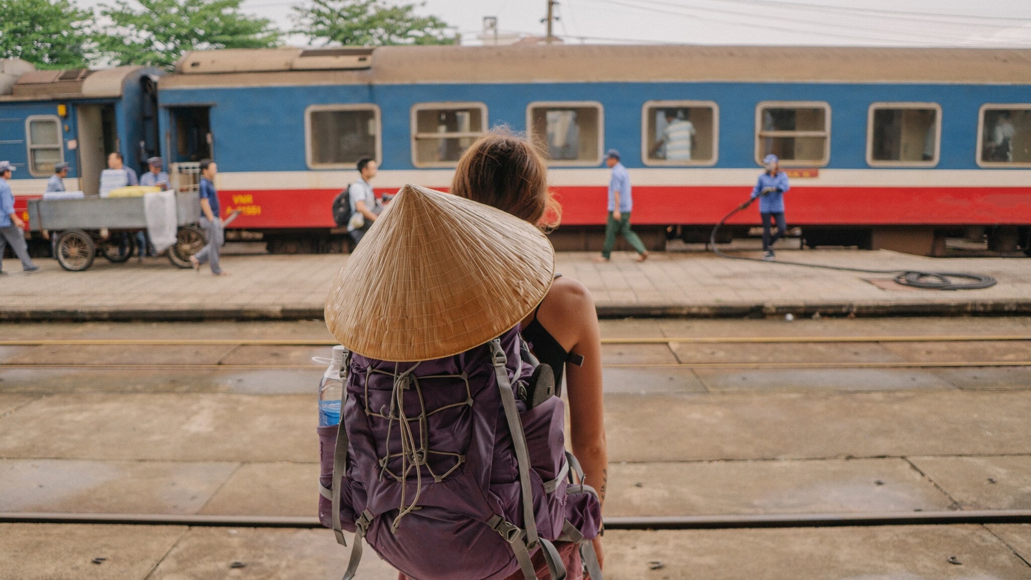 Eine Person mit einem großen, runden Strohhut und einem lila Rucksack steht auf einem Bahnsteig und schaut auf einen Zug. Im Hintergrund sind mehrere Personen und ein Zug zu sehen. Eine Person mit einem großen, runden Strohhut und einem lila Rucksack steht auf einem Bahnsteig und schaut auf einen Zug. Im Hintergrund sind mehrere Personen und ein Zug zu sehen.