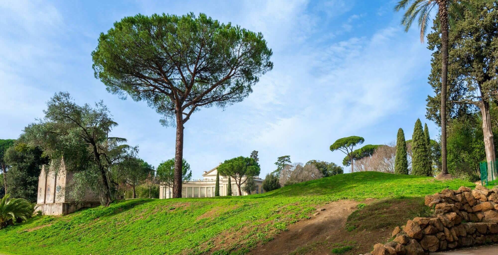 Grüne Wiese mit Treppe und Steinmauer im Villa Torlonia Park in Rom, umgeben von hohen Pinien und Zypressen unter blauem Himmel.
