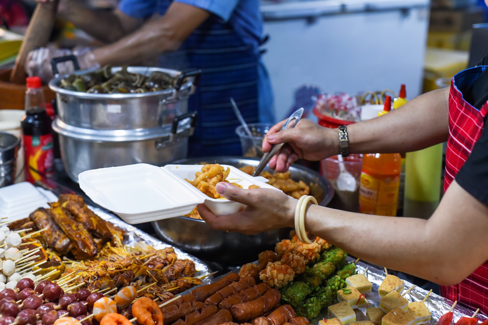 Eine Person gibt Essen zum Mitnehmen in eine Box, vor ihr liegen verschiedene Spieße.