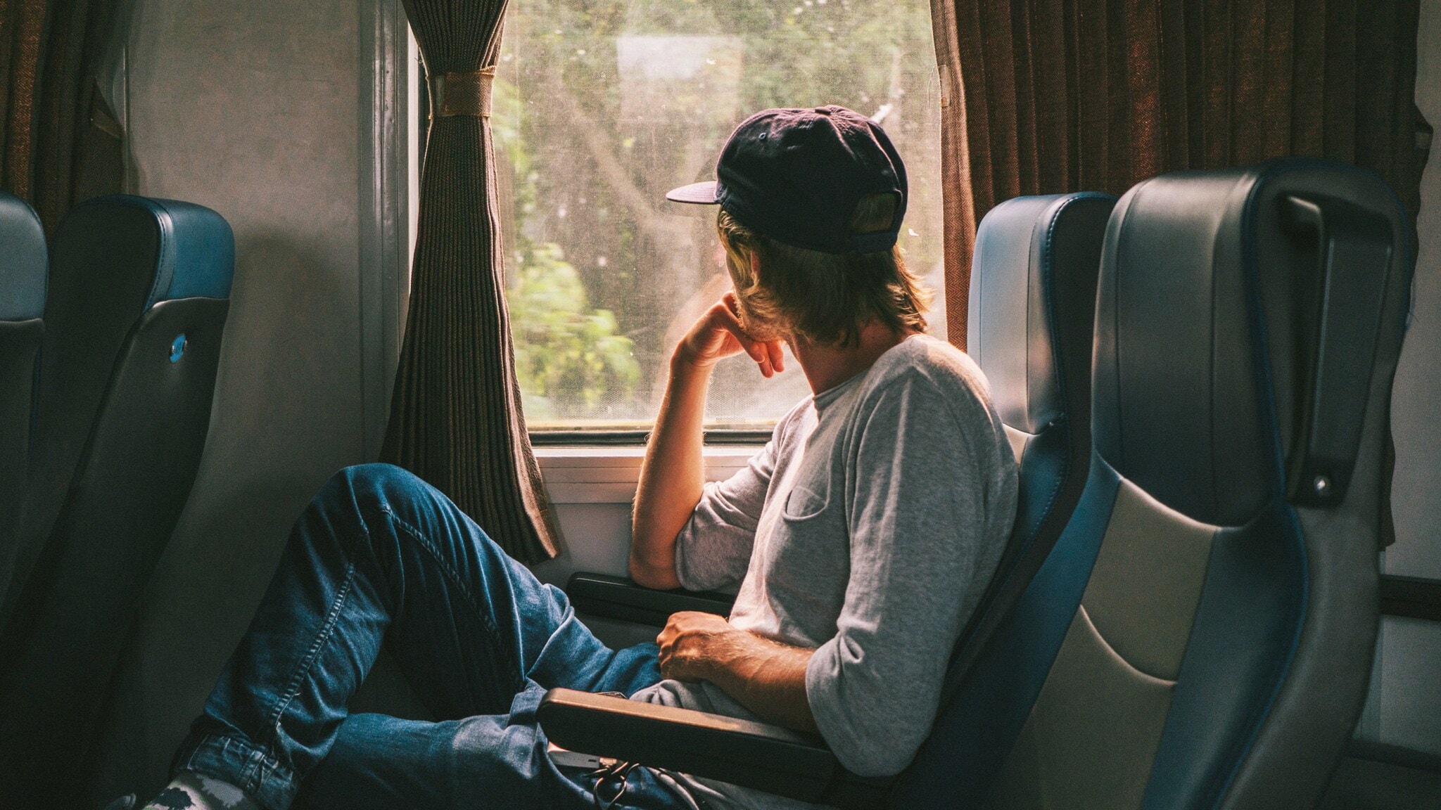 Ein Mann mit einer Baseballmütze sitzt in einem Zug und schaut nachdenklich aus dem Fenster. Er trägt ein graues Oberteil und Jeans. Ein Mann mit einer Baseballmütze sitzt in einem Zug und schaut nachdenklich aus dem Fenster. Er trägt ein graues Oberteil und Jeans.