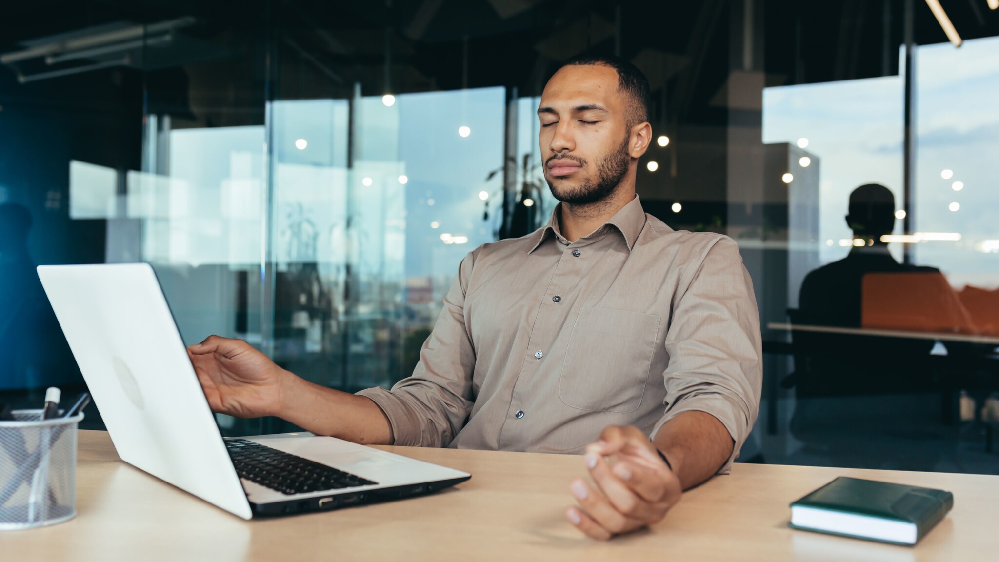 Ein Mann sitzt mit geschlossenen Augen in Meditationshaltung vor einem Laptop an einem Schreibtisch in einem Büro.