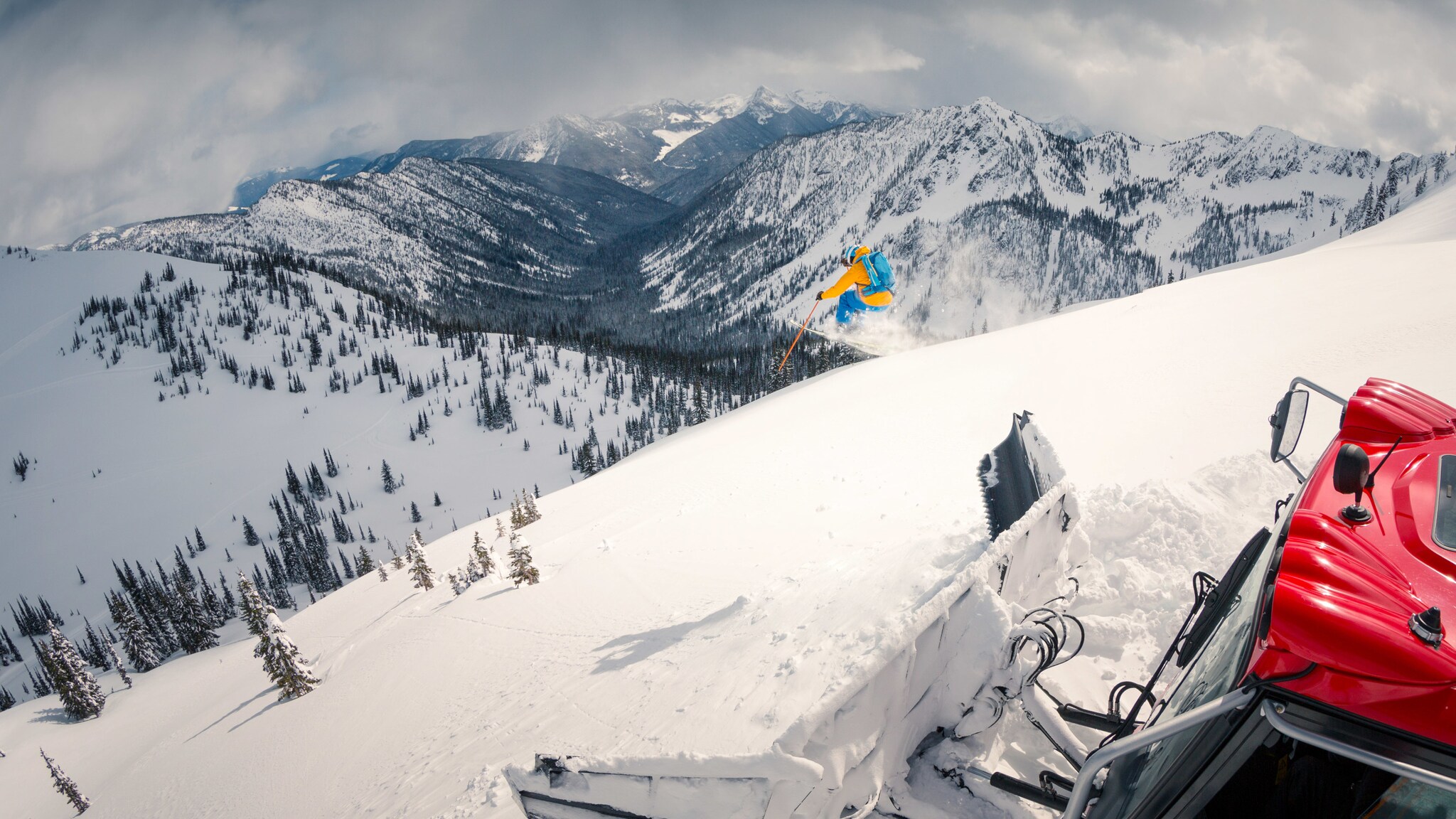 Ein Skifahrer springt einen Schneehang hinunter vor Bergpanorama, im Vordergrund eine Pistenplanierraupe im Tiefschnee.