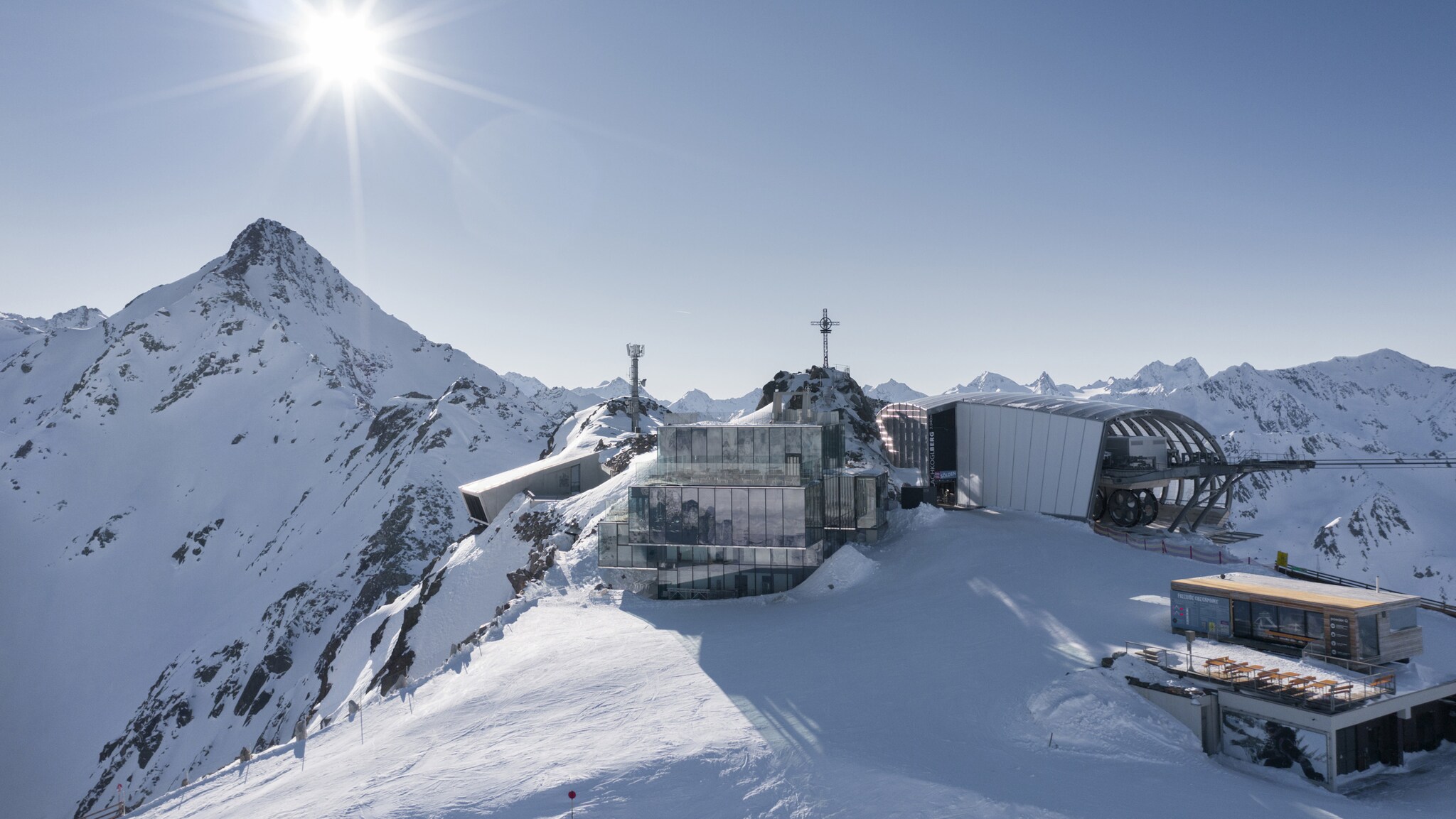 Kubistischer Gebäudekomplex mit verspiegelter Fassade neben einer Seilbahnstation auf einem schneebedeckten Gipfel bei Sonnenschein. Kubistischer Gebäudekomplex mit verspiegelter Fassade neben einer Seilbahnstation auf einem schneebedeckten Gipfel bei Sonnenschein.