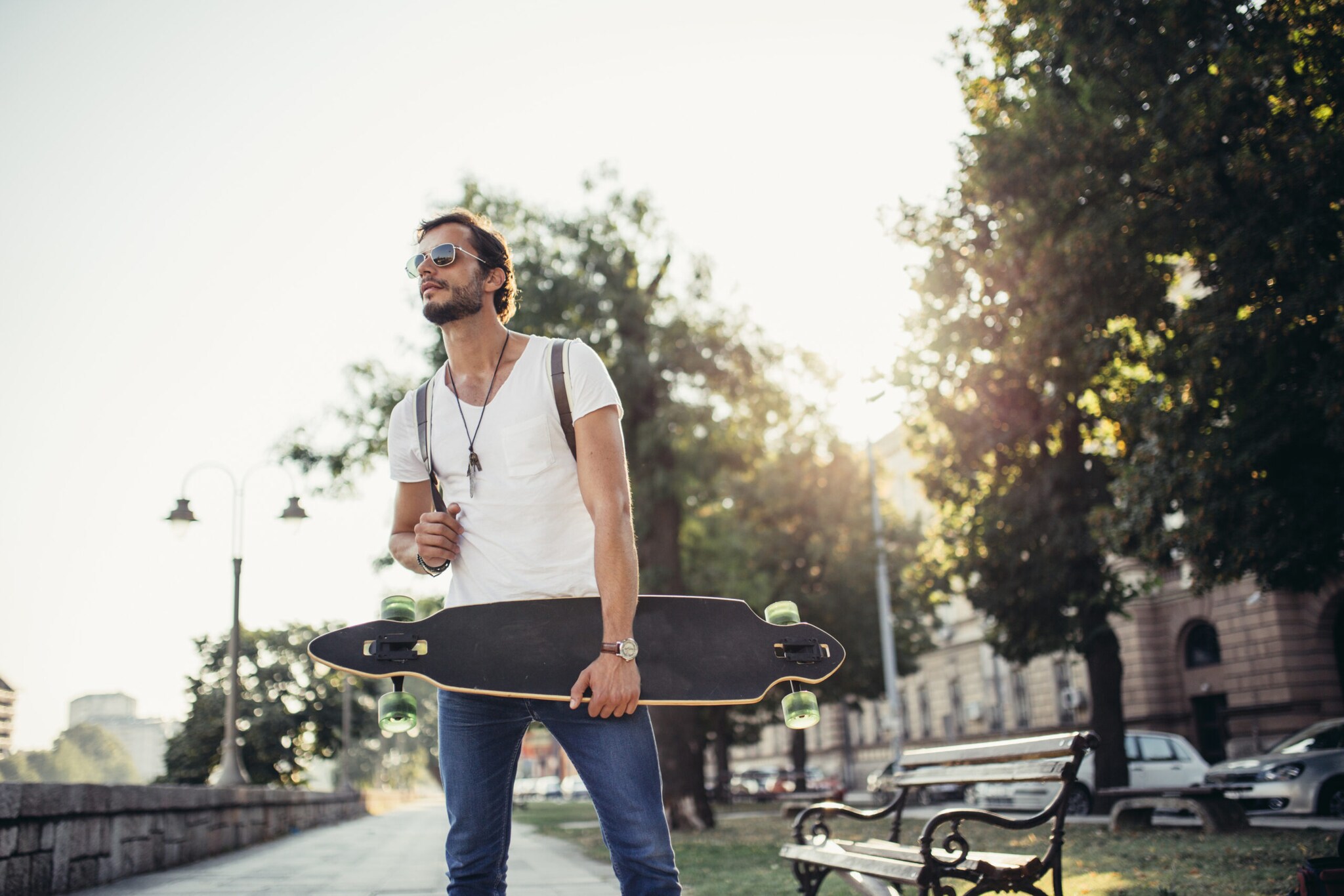 Mann mit Sonnenbrille hält ein Longboard, dahinter Stadtkulisse