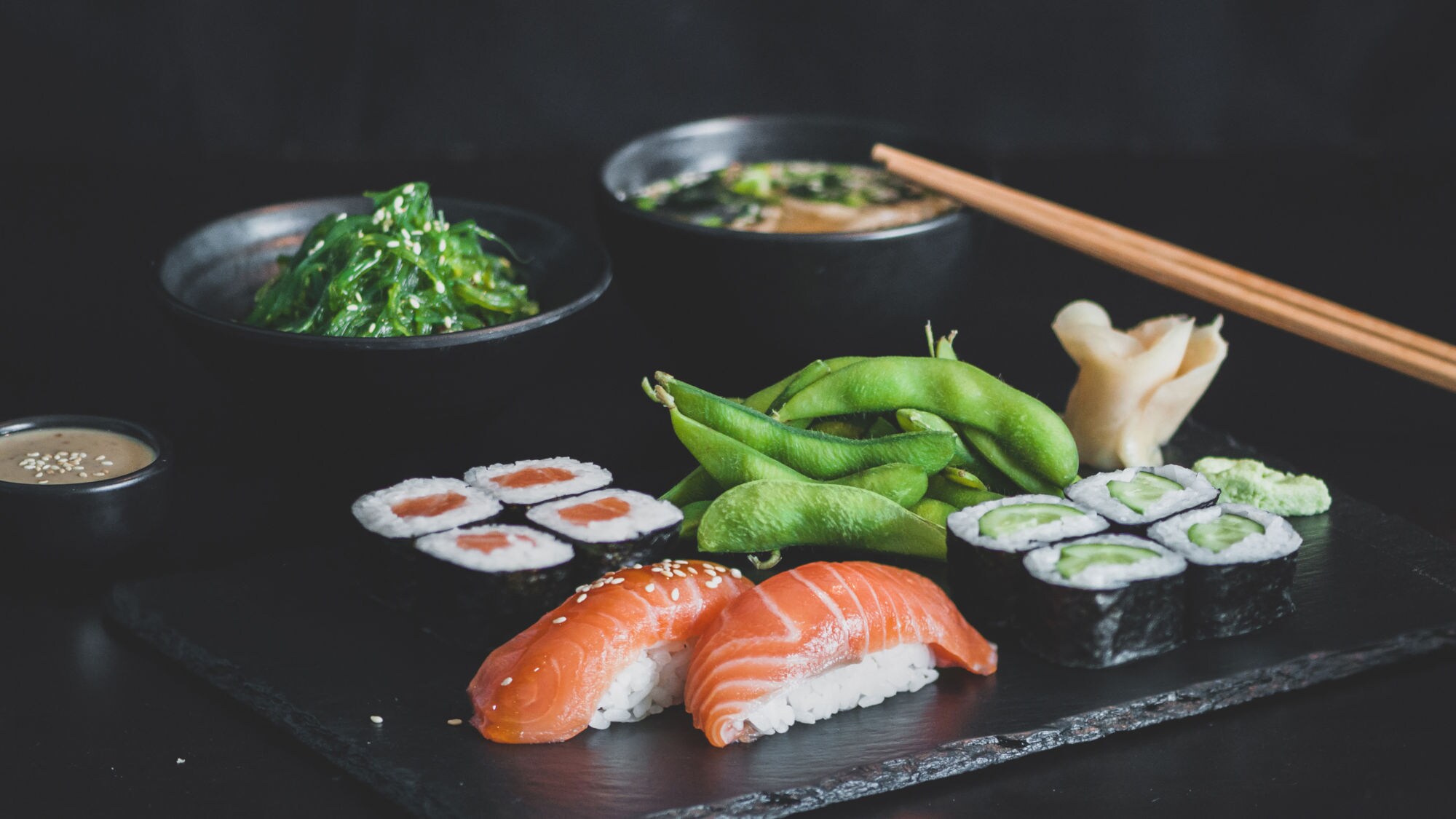 Ein schwarzes Servierbrett mit Sushi, darunter zwei Lachs-Sashimi-Stücke, eine Rolle mit Lachs und Avocado, Edamame, Algensalat, eingelegtem Ingwer und Miso-Suppe in Schalen. Essstäbchen liegen daneben.