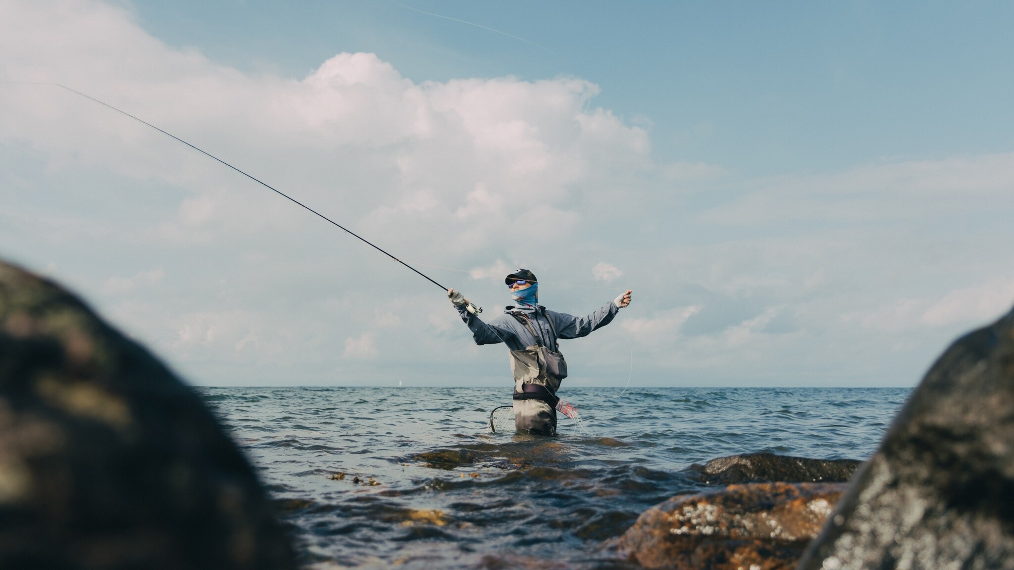 Angler steht hüfttief im Wasser zwischen Felsen und wirft eine Angelrute aus, bewölkter Himmel im Hintergrund. Angler steht hüfttief im Wasser zwischen Felsen und wirft eine Angelrute aus, bewölkter Himmel im Hintergrund.