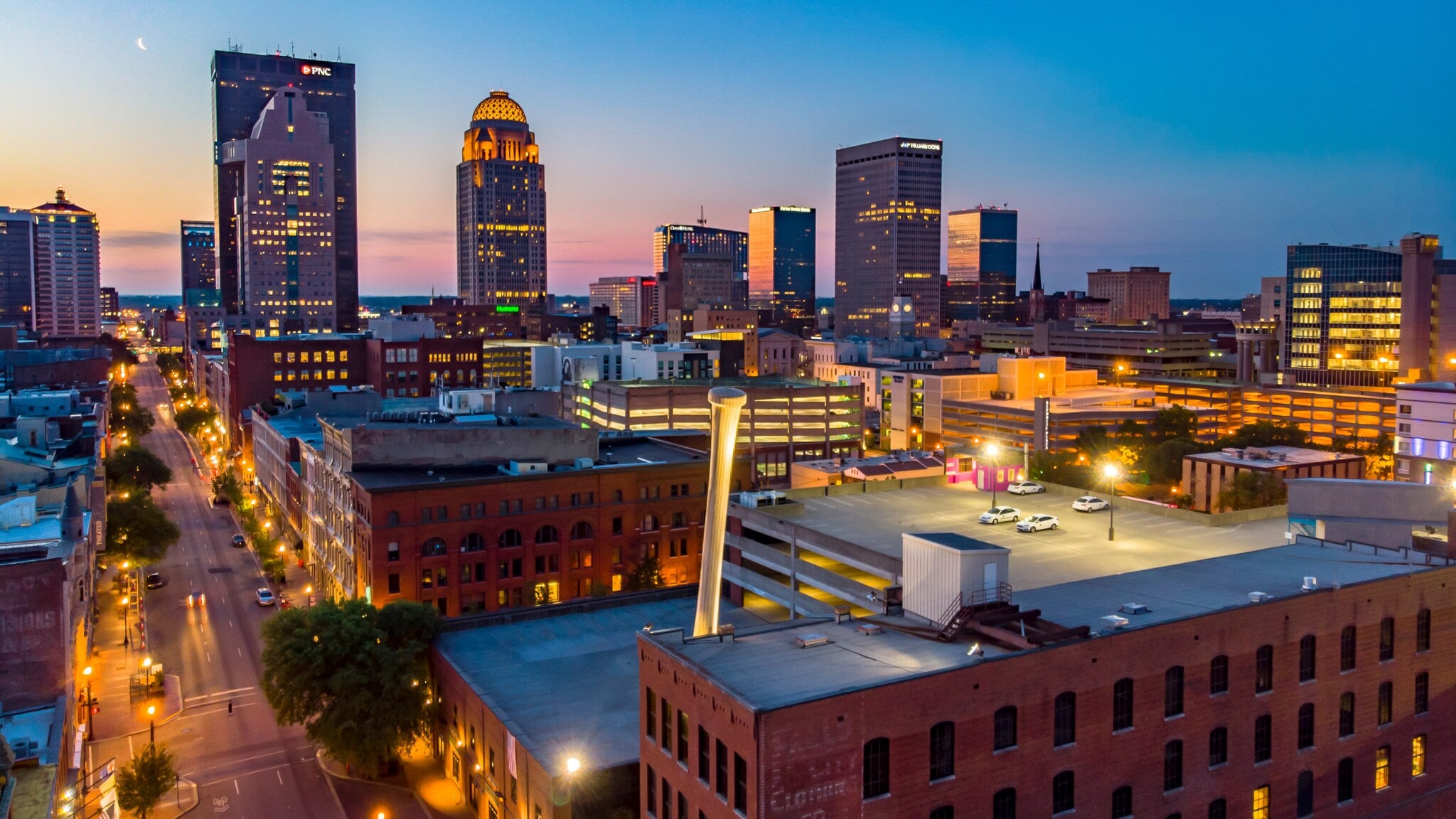 Skyline von Louisville, Kentucky, bei Sonnenuntergang mit leuchtenden Hochhäusern, historischen Backsteingebäuden im Vordergrund und der beleuchteten Innenstadtstraße. Skyline von Louisville, Kentucky, bei Sonnenuntergang mit leuchtenden Hochhäusern, historischen Backsteingebäuden im Vordergrund und der beleuchteten Innenstadtstraße.