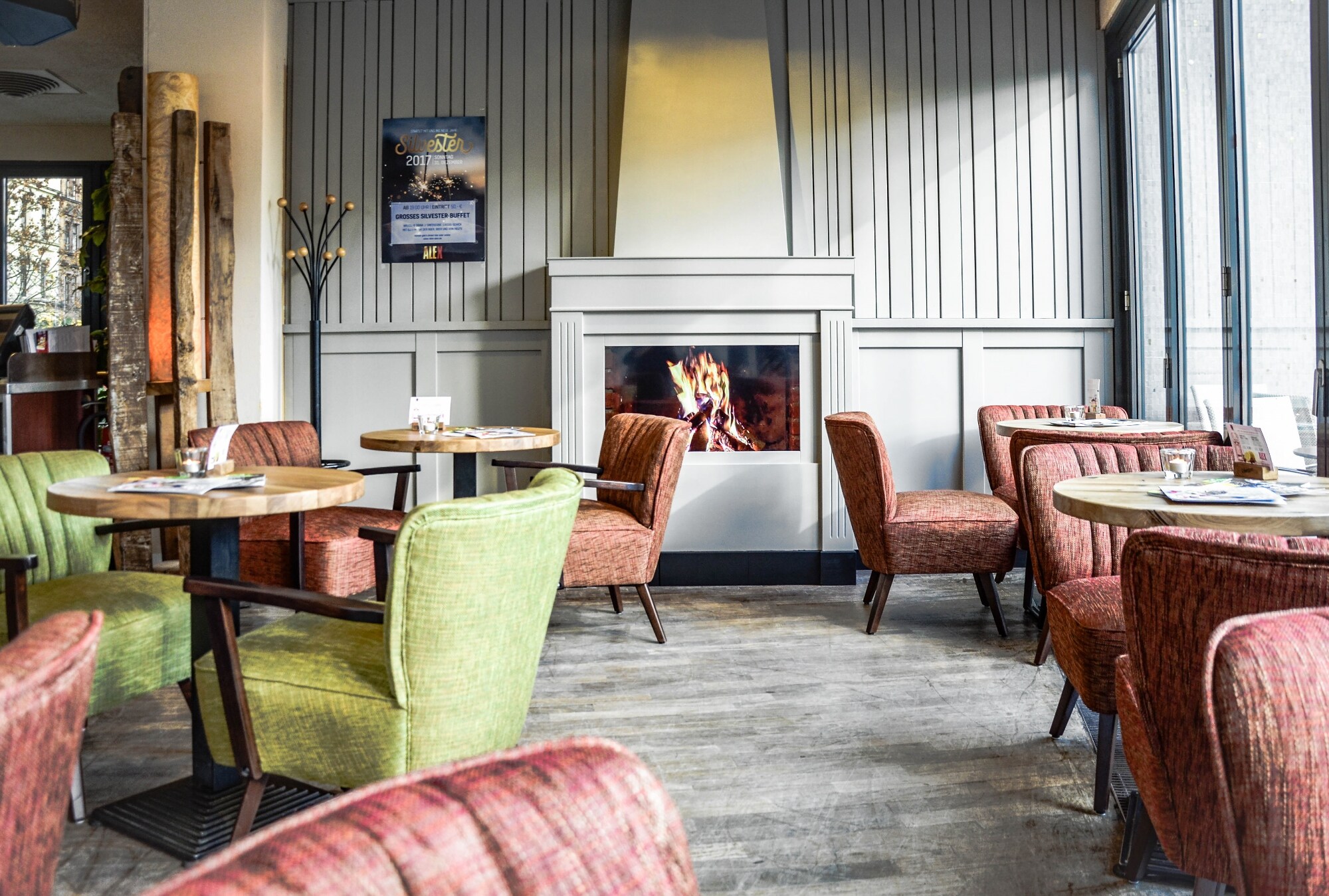 Gemütlicher Gastraum des ALEX in Wiesbaden mit loderndem Gaskamin, runden kleinen Tischen und bequemen Sesseln in Grün und Rot.