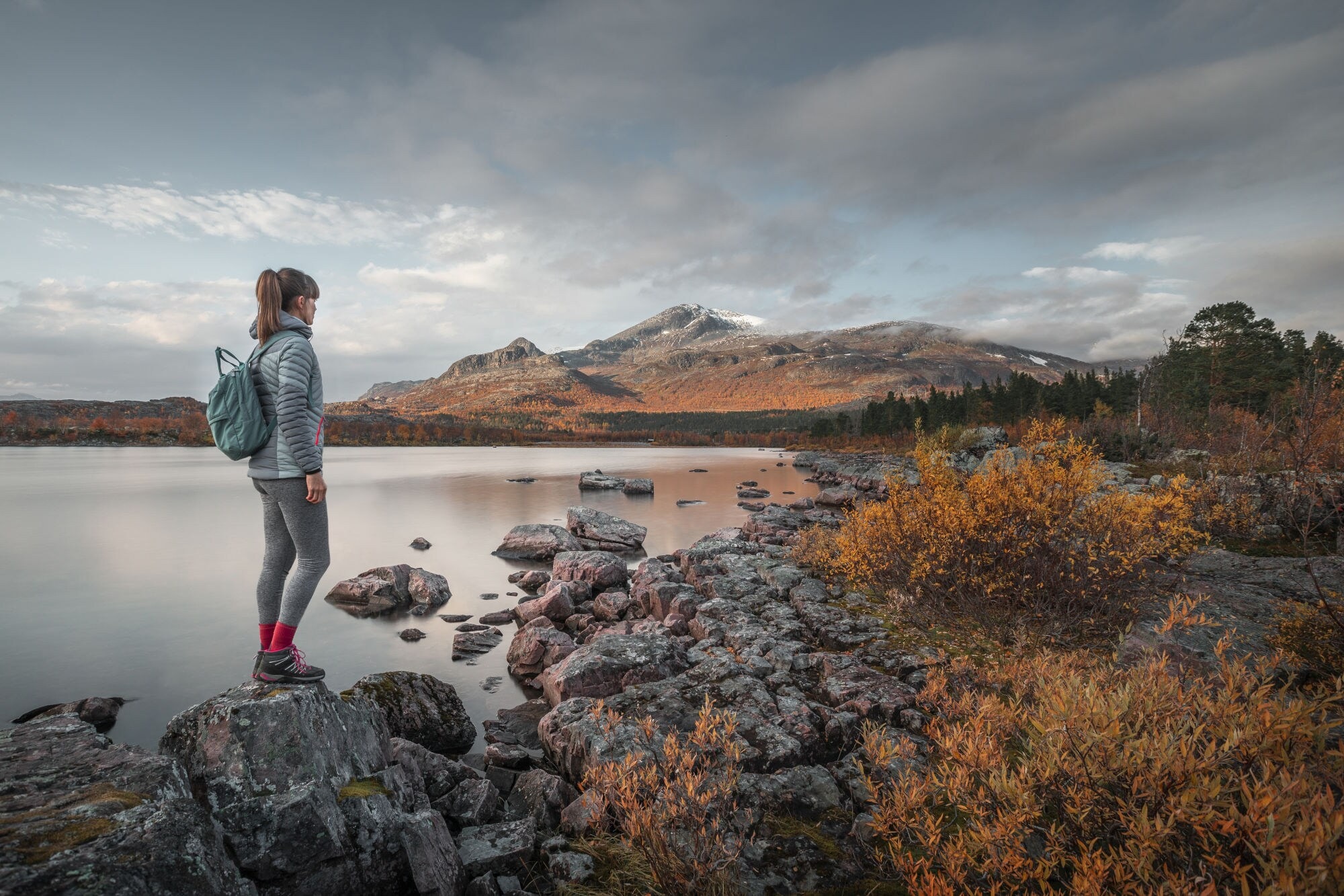 Eine Frau mit Rucksack steht auf einem Stein an einem See in flacher Felslandschaft mit herbstlicher Vegetation. Eine Frau mit Rucksack steht auf einem Stein an einem See in flacher Felslandschaft mit herbstlicher Vegetation.
