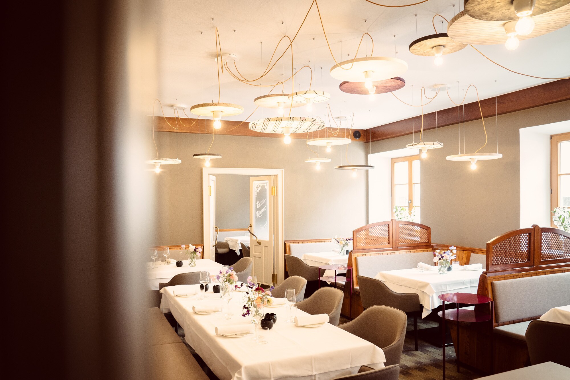 Der Speiseraum eines modern gestalteten Wirtshauses mit weiß eingedeckten Tischen, gepolsterten Stühlen und vielen runden Deckenlampen