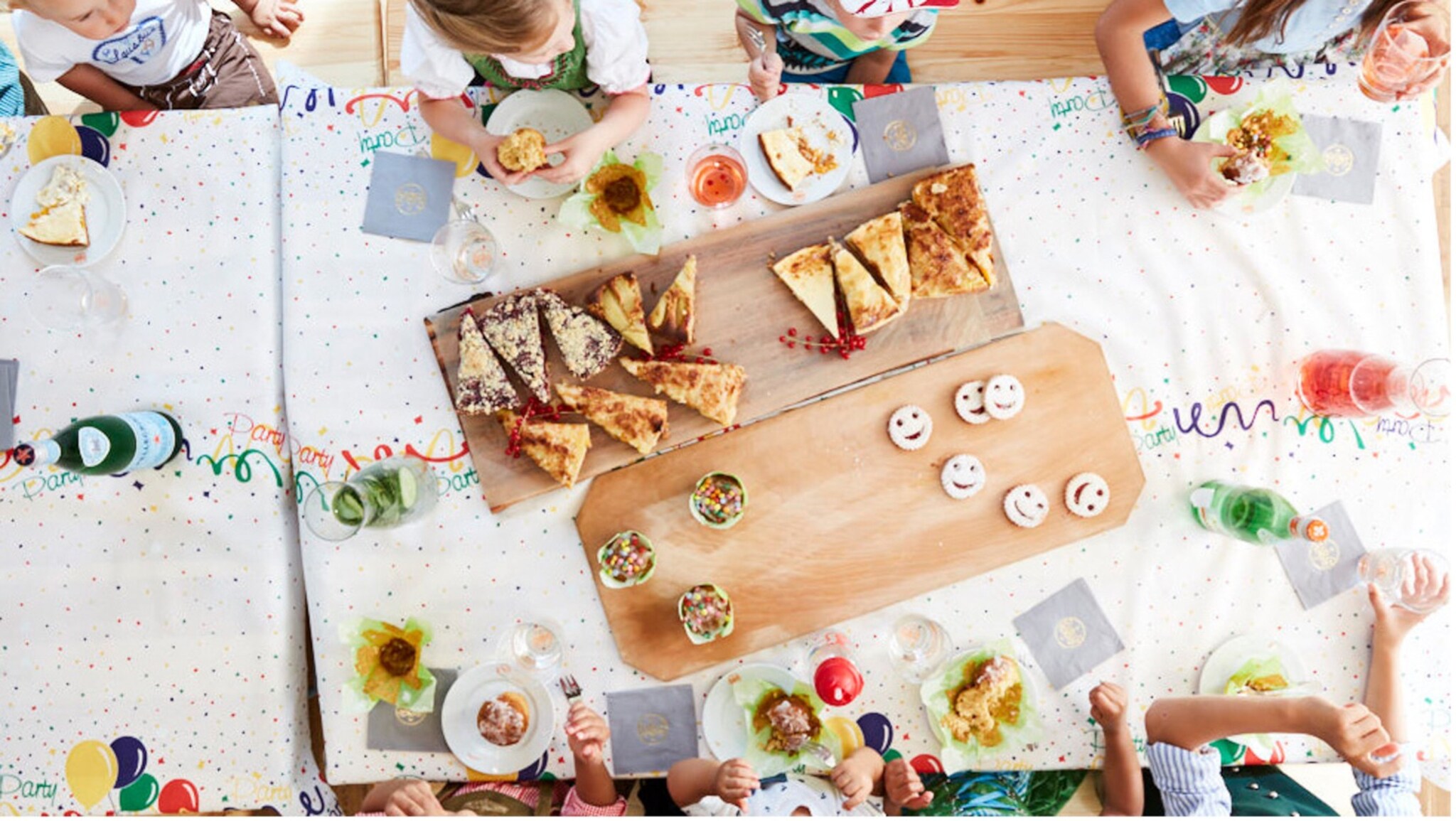 Kinder sitzen an einem Tisch mit buntem Tischtuch und essen Pizza und Kekse mit lachenden Gesichtern