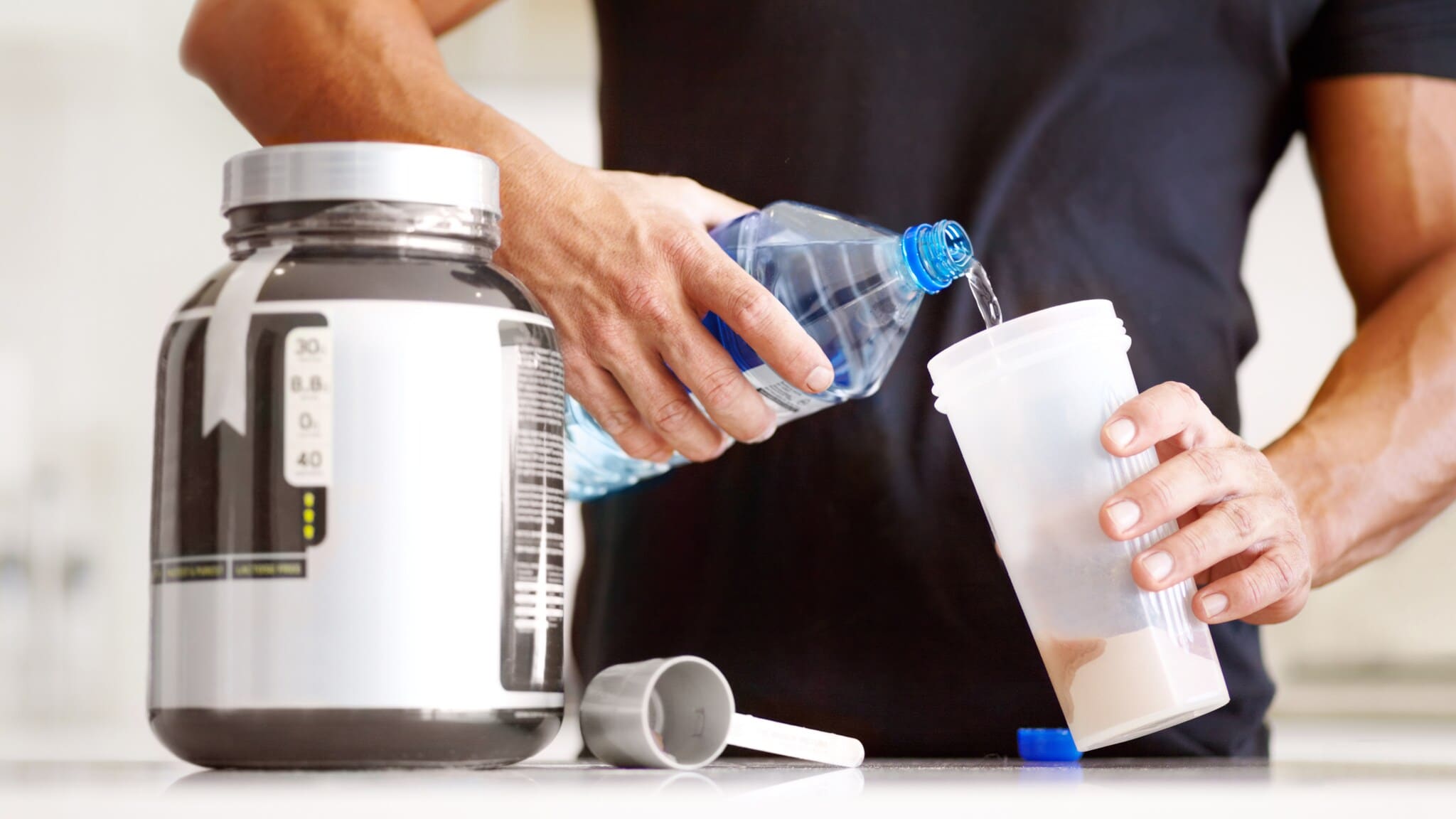 Mann gießt Wasser aus einer Flasche in einen Protein-Shaker neben einem großen Behälter mit Proteinpulver Mann gießt Wasser aus einer Flasche in einen Protein-Shaker neben einem großen Behälter mit Proteinpulver