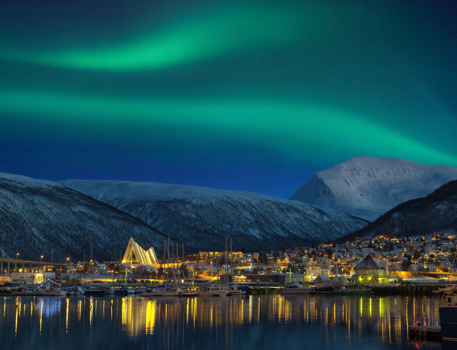Eine beleuchtete Stadt bei Nacht vor schneebedeckten Bergen und grün schimmernden Polarlichtern am Himmel Eine beleuchtete Stadt bei Nacht vor schneebedeckten Bergen und grün schimmernden Polarlichtern am Himmel
