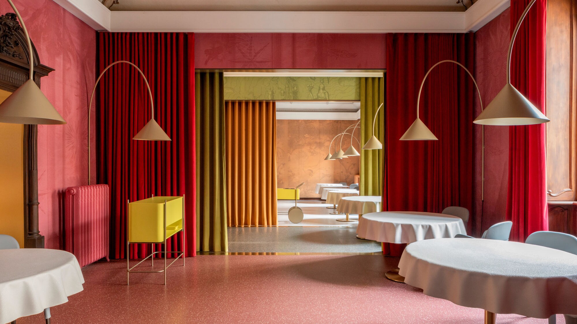 Ein eleganter Innenraum mit runden Tischen, die mit weißen Tischdecken bedeckt sind. An den Wänden hängen rote und grüne Vorhänge. Im Hintergrund sind gelbe und orangefarbene Wände sichtbar. Ein eleganter Innenraum mit runden Tischen, die mit weißen Tischdecken bedeckt sind. An den Wänden hängen rote und grüne Vorhänge. Im Hintergrund sind gelbe und orangefarbene Wände sichtbar.