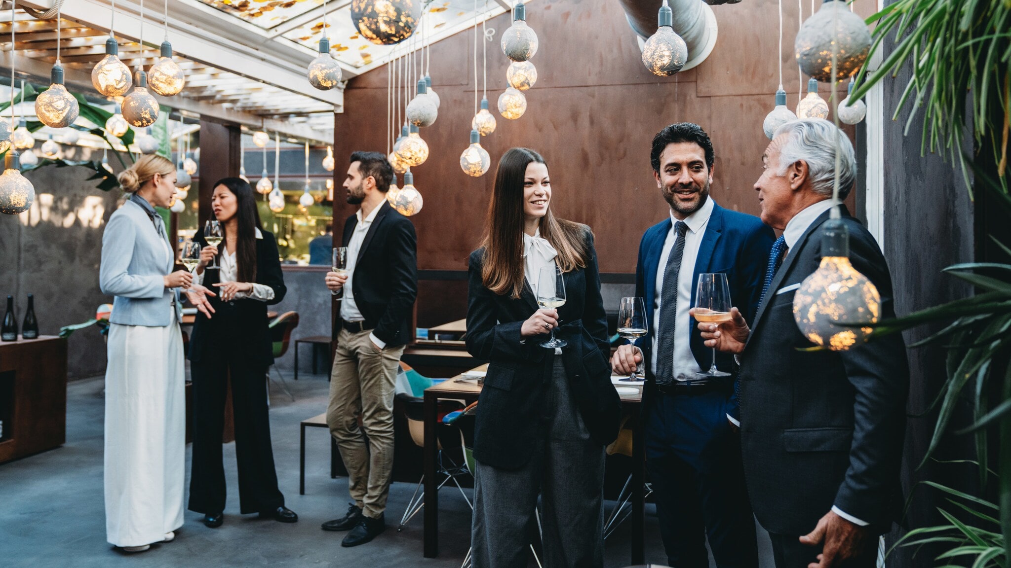 Personen in eleganter Kleidung stehen mit Weingläsern in den Händen beieinander in einem elegant geschmückten Raum. Personen in eleganter Kleidung stehen mit Weingläsern in den Händen beieinander in einem elegant geschmückten Raum.