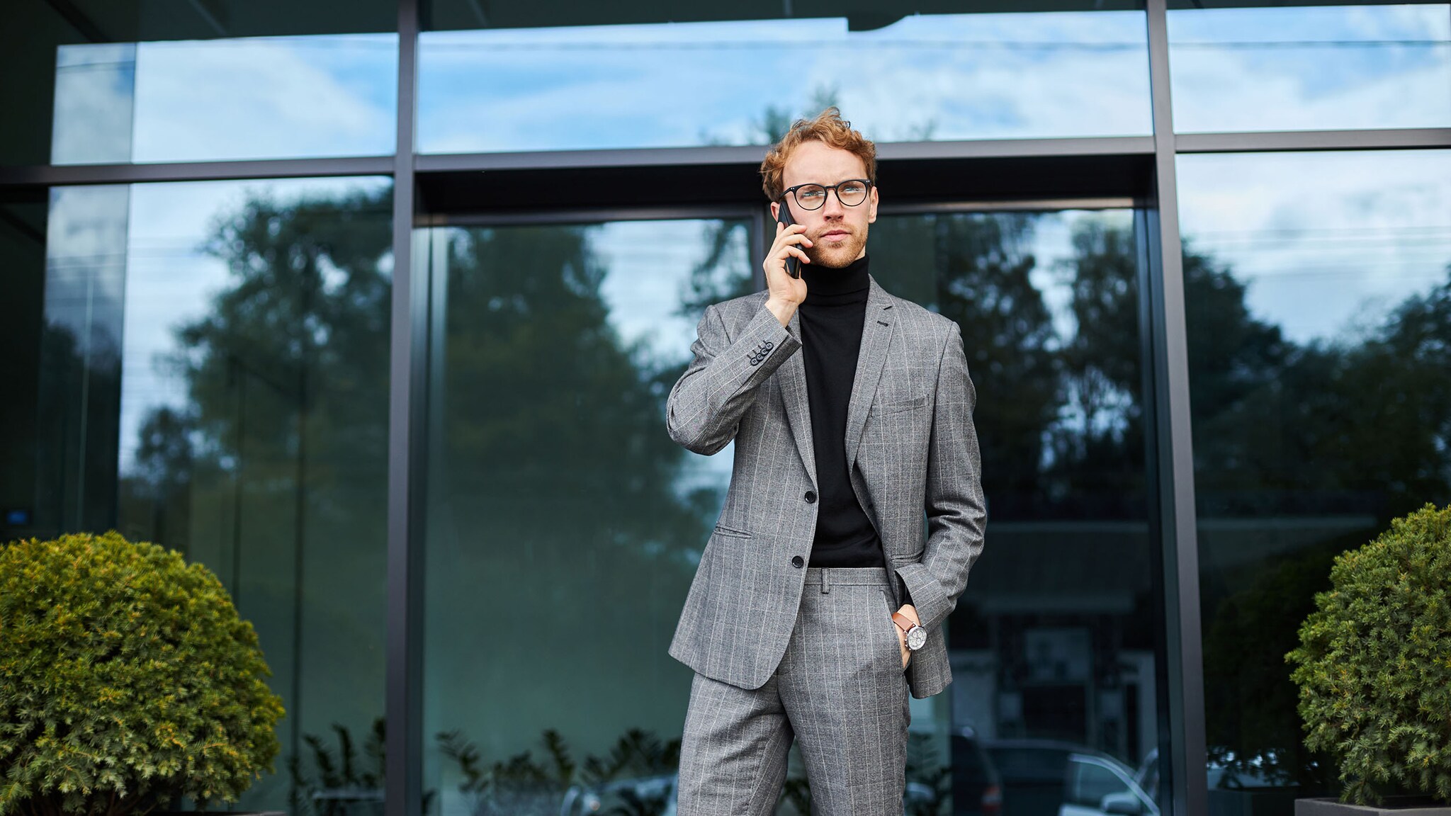 Ein stilvoll gekleideter Mann im grauen Anzug und schwarzen Rollkragenpullover telefoniert mit Smartphone vor einem Gebäude.