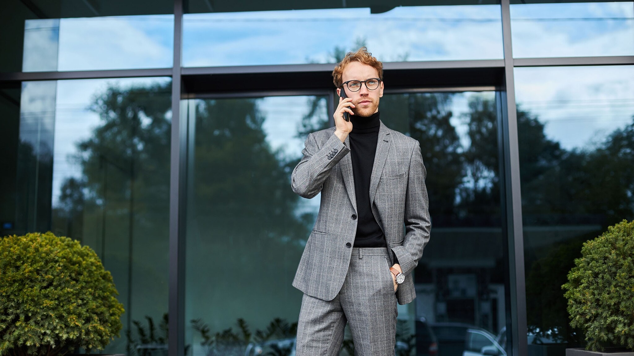 Businessman having phone call outdoors near office building Ein stilvoll gekleideter Mann im grauen Anzug und schwarzen Rollkragenpullover telefoniert mit Smartphone vor einem Gebäude.