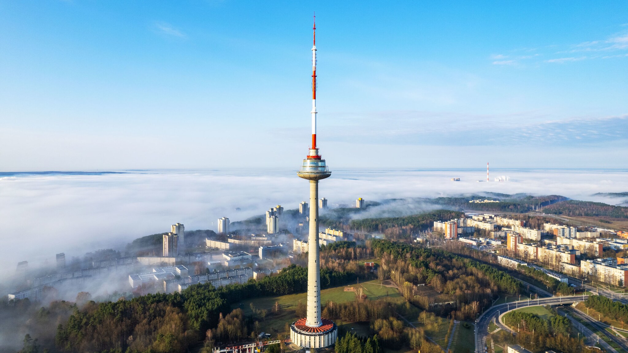 Hoher Fernsehturm mit rot-weiß gestreifter Spitze vor einem nebelverhangenen Stadtpanorama.