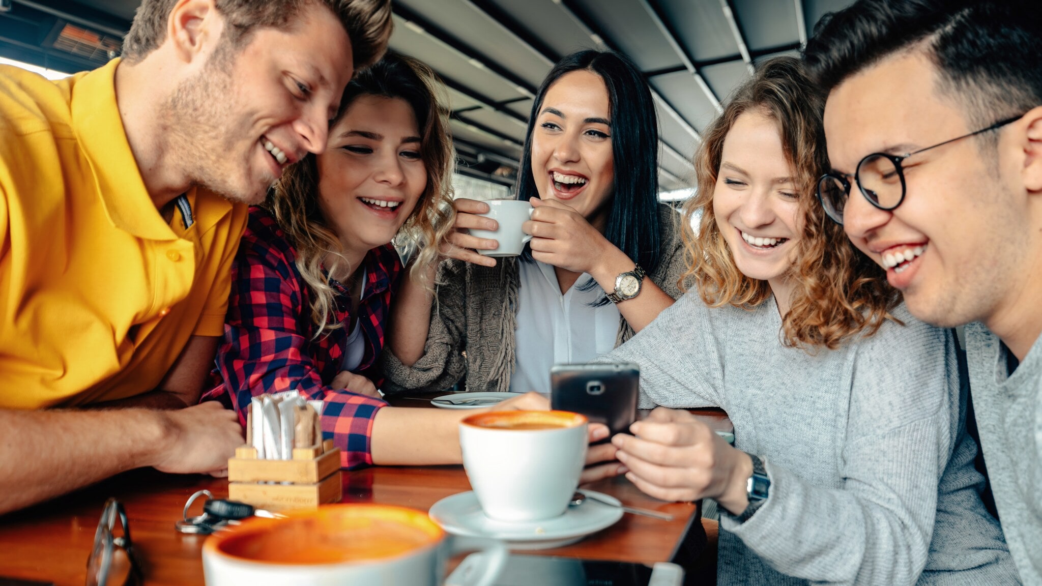 Fünf junge Erwachsene sitzen gut gelaunt an einem Tisch mit Kaffeetassen und schauen gemeinsam auf ein Smartphone. Fünf junge Erwachsene sitzen gut gelaunt an einem Tisch mit Kaffeetassen und schauen gemeinsam auf ein Smartphone.