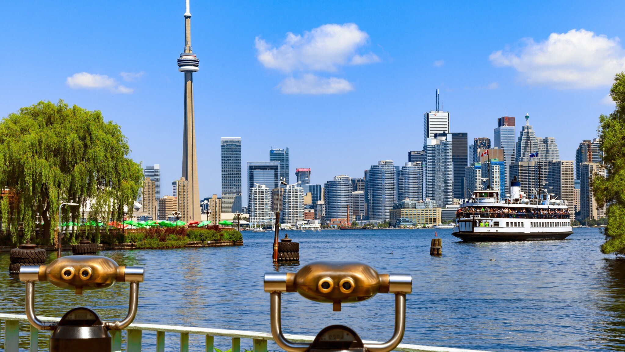 Skyline von Toronto am See mit Fähre zwischen begrünten Ufern, im Vordergrund zwei Aussichtsfernrohre aus Metall.
