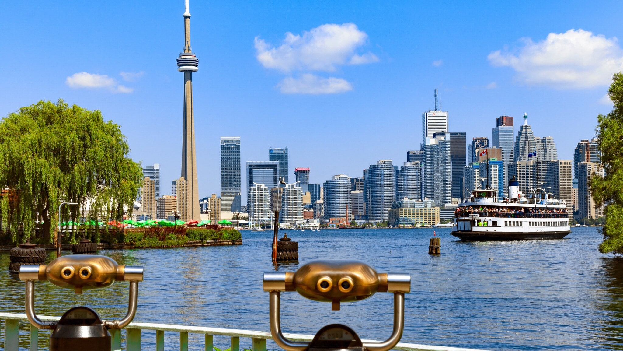 Skyline von Toronto am See mit Fähre zwischen begrünten Ufern, im Vordergrund zwei Aussichtsfernrohre aus Metall. Skyline von Toronto am See mit Fähre zwischen begrünten Ufern, im Vordergrund zwei Aussichtsfernrohre aus Metall.