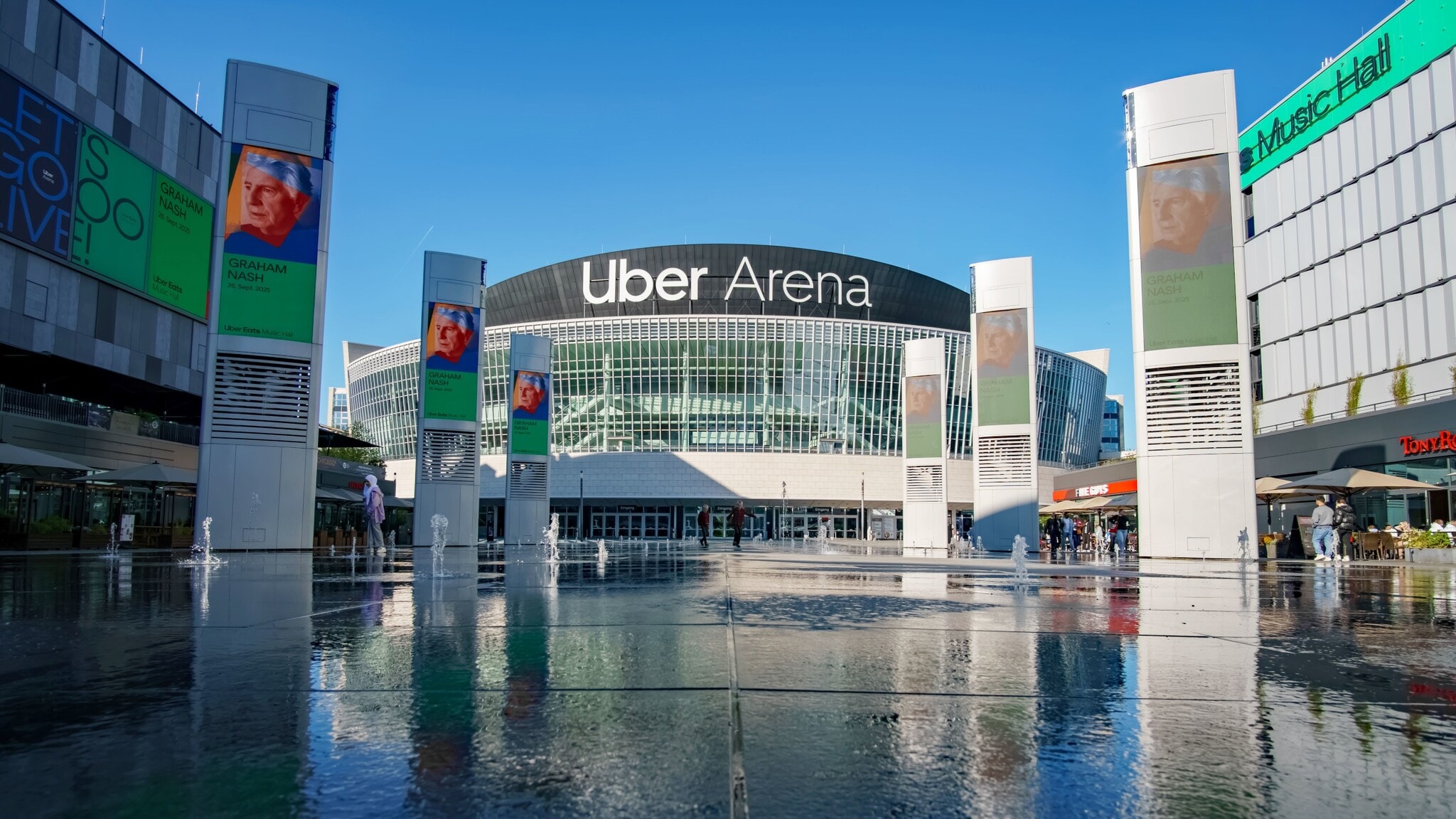 Rundes Gebäude mit Glasfassade und der Aufschrift Uber Arena hinter einem bebauten Platz mit Werbetafeln um eine Wasserfläche. Rundes Gebäude mit Glasfassade und der Aufschrift Uber Arena hinter einem bebauten Platz mit Werbetafeln um eine Wasserfläche.