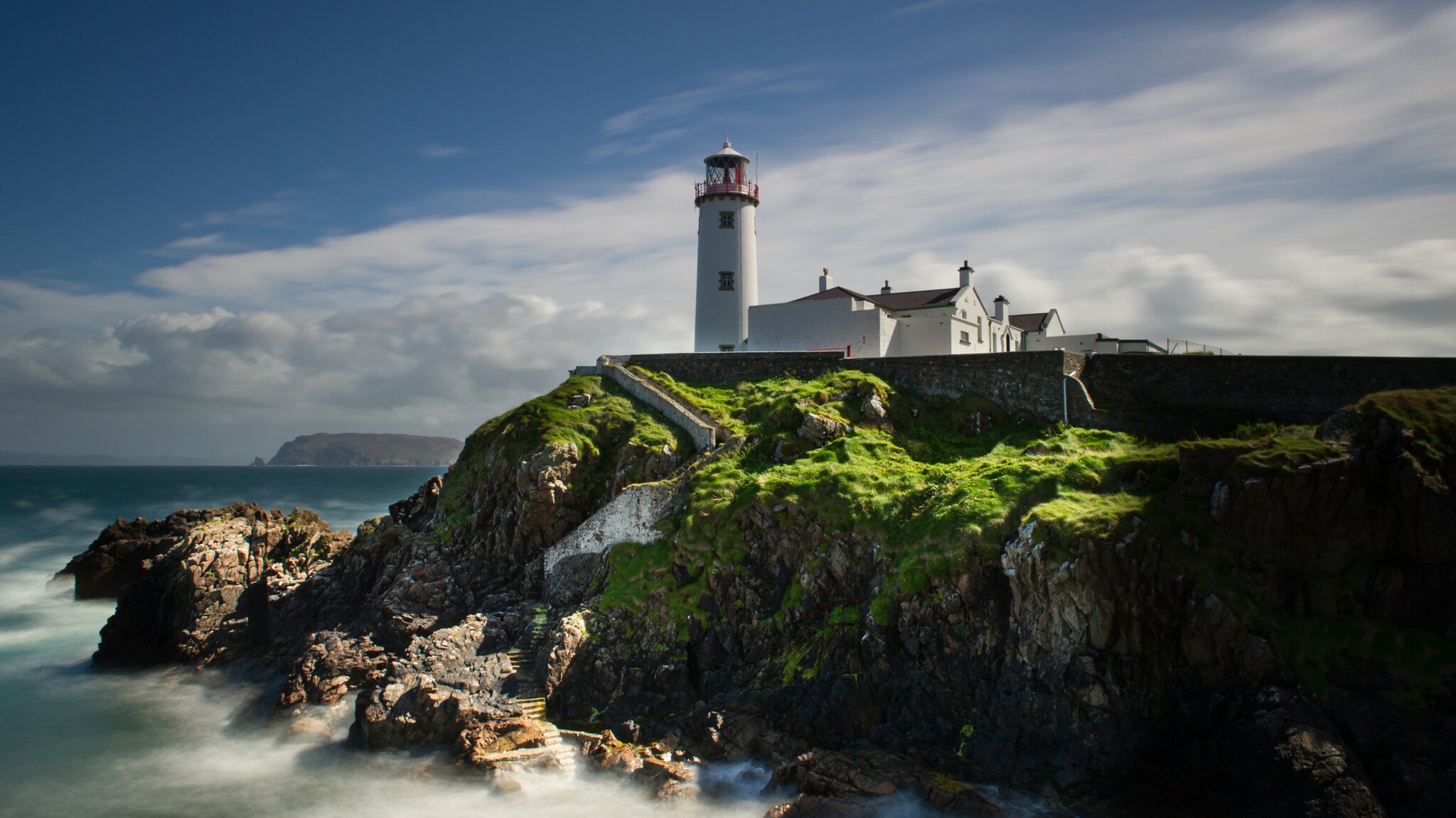 Ein Leuchtturm mit rotem Licht auf einem Hügel, umgeben von grünem Gras und steiniger Küste, mit Wellen, die gegen die Felsen schlagen. Ein Leuchtturm mit rotem Licht auf einem Hügel, umgeben von grünem Gras und steiniger Küste, mit Wellen, die gegen die Felsen schlagen.