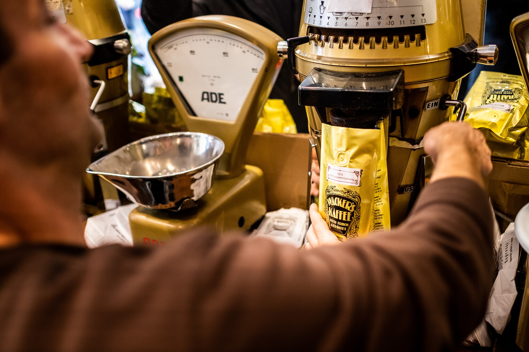 Eine Person von hinten in einem Kaffeegeschäft vor einer mechanischen Waage bei der Abfüllung von Bohnen in eine gelbe Verpackung