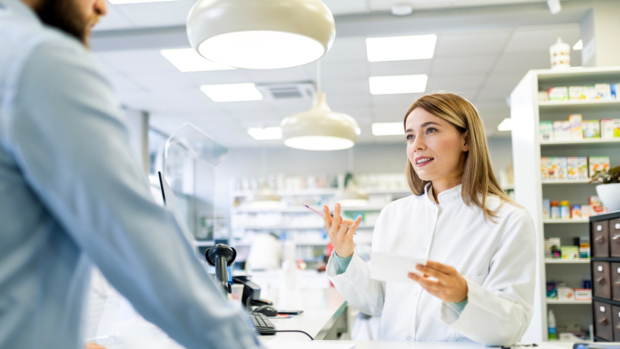 Apothekerin in weißem Kittel berät Kunden an der Apothekentheke in moderner Apotheke.