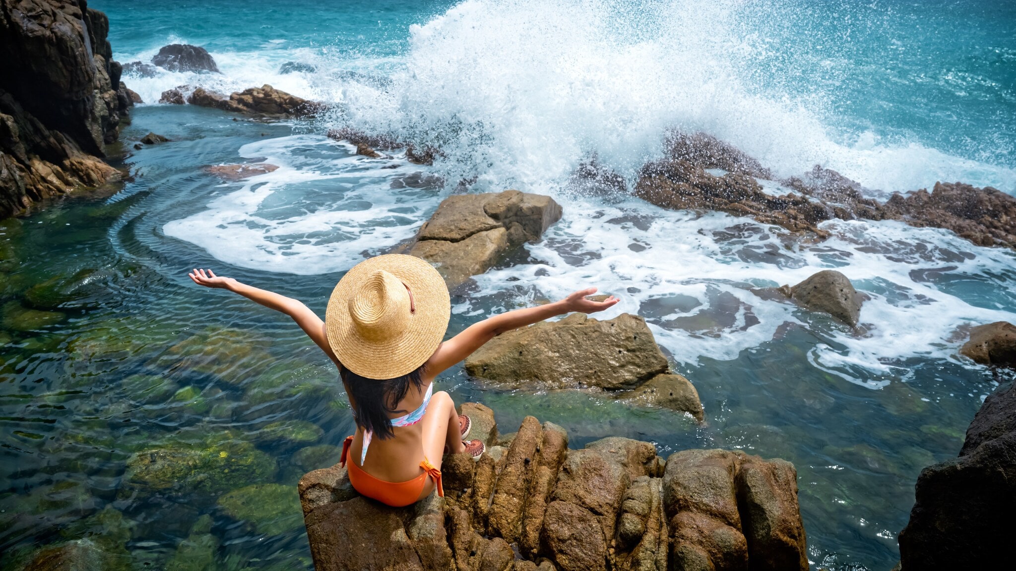 Frau mit Strohhut und orangefarbenem Bikini sitzt auf Felsen am Meer, brechende Wellen und bewölkter Himmel im Hintergrund Frau mit Strohhut und orangefarbenem Bikini sitzt auf Felsen am Meer, brechende Wellen und bewölkter Himmel im Hintergrund