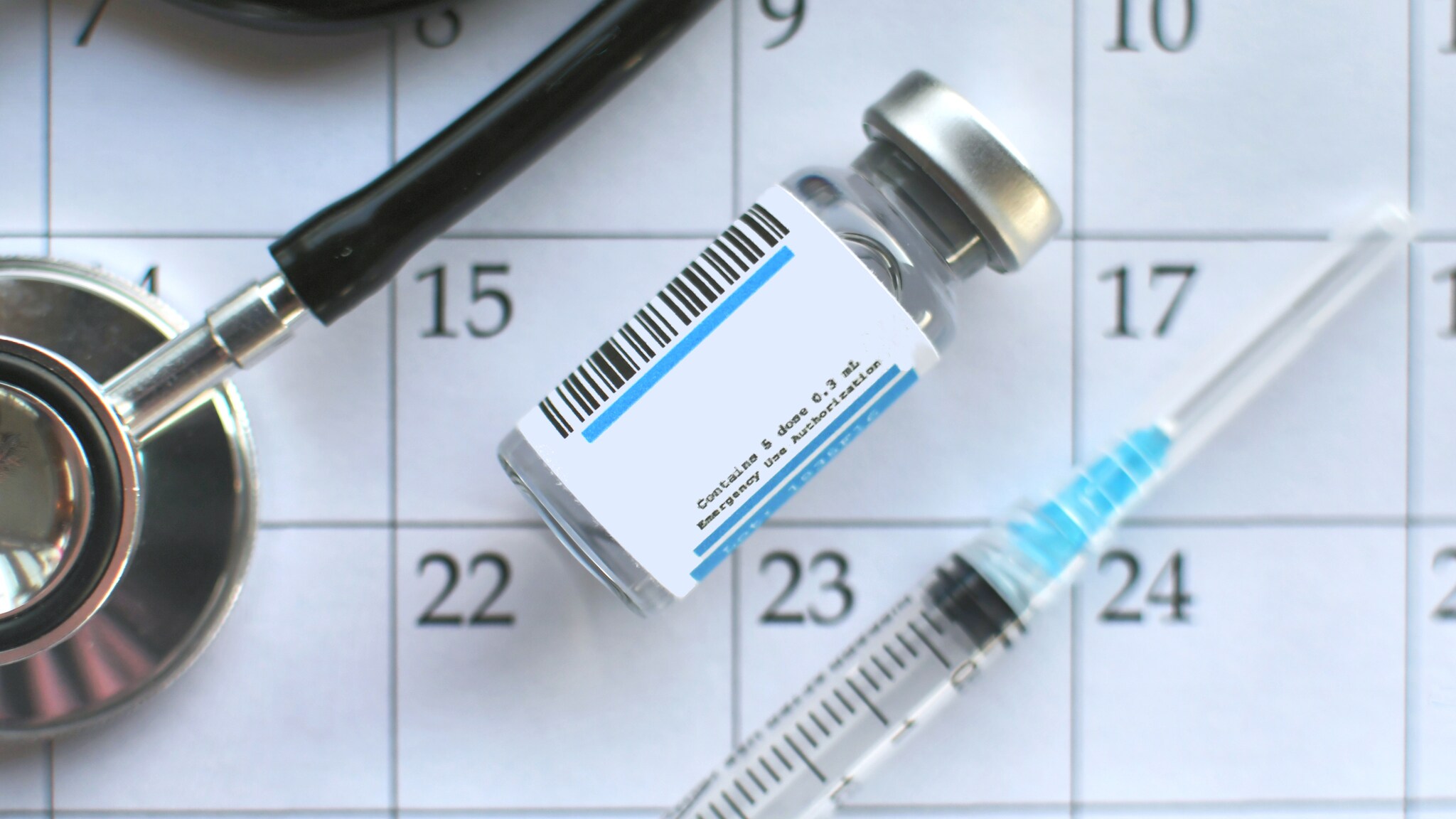 Impfstofffläschchen mit Barcode, Spritze und Stethoskop auf einem Kalenderblatt mit den Tagen 15 bis 23 sichtbar