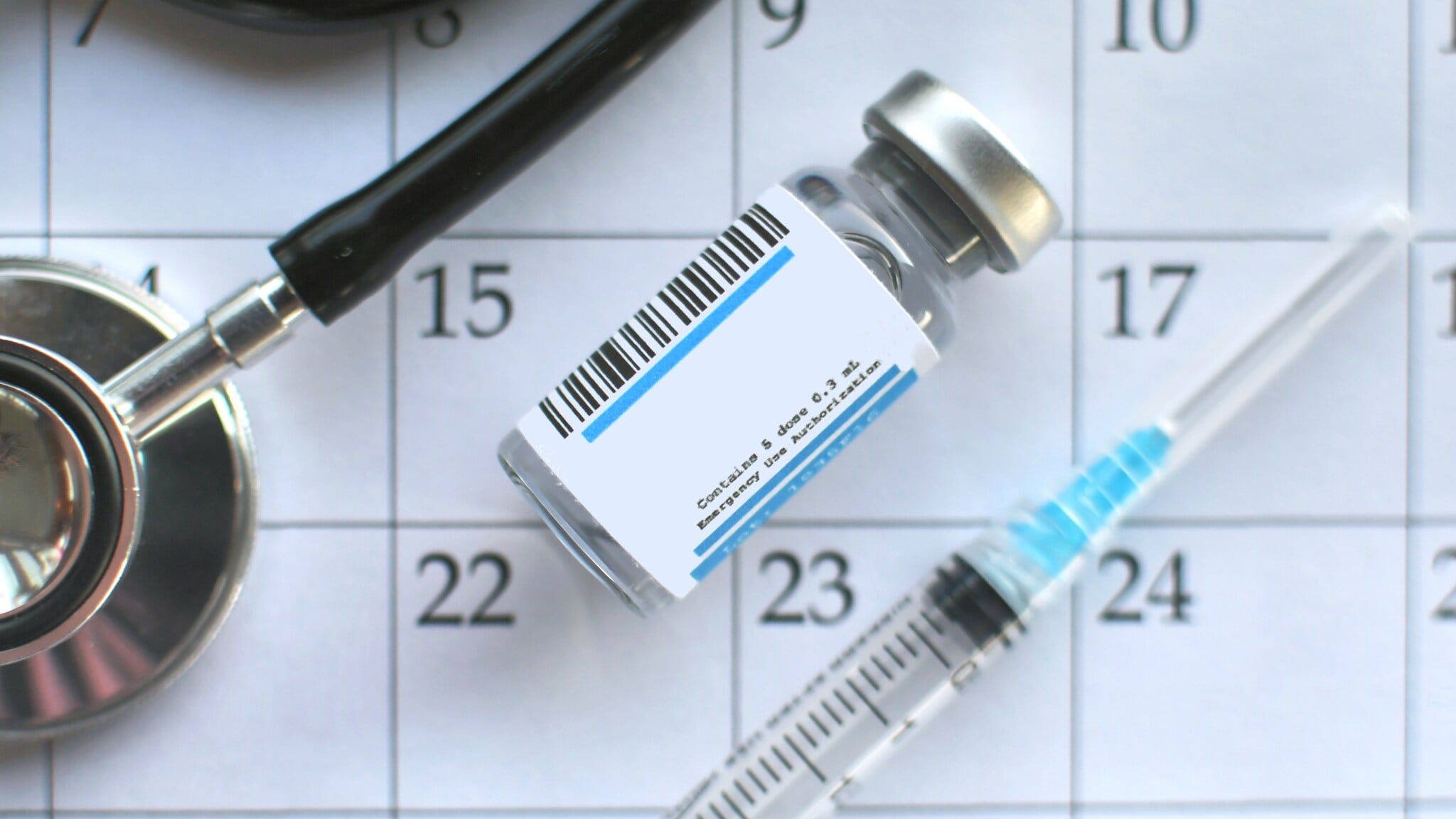 Impfstofffläschchen mit Barcode, Spritze und Stethoskop auf einem Kalenderblatt mit den Tagen 15 bis 23 sichtbar Impfstofffläschchen mit Barcode, Spritze und Stethoskop auf einem Kalenderblatt mit den Tagen 15 bis 23 sichtbar