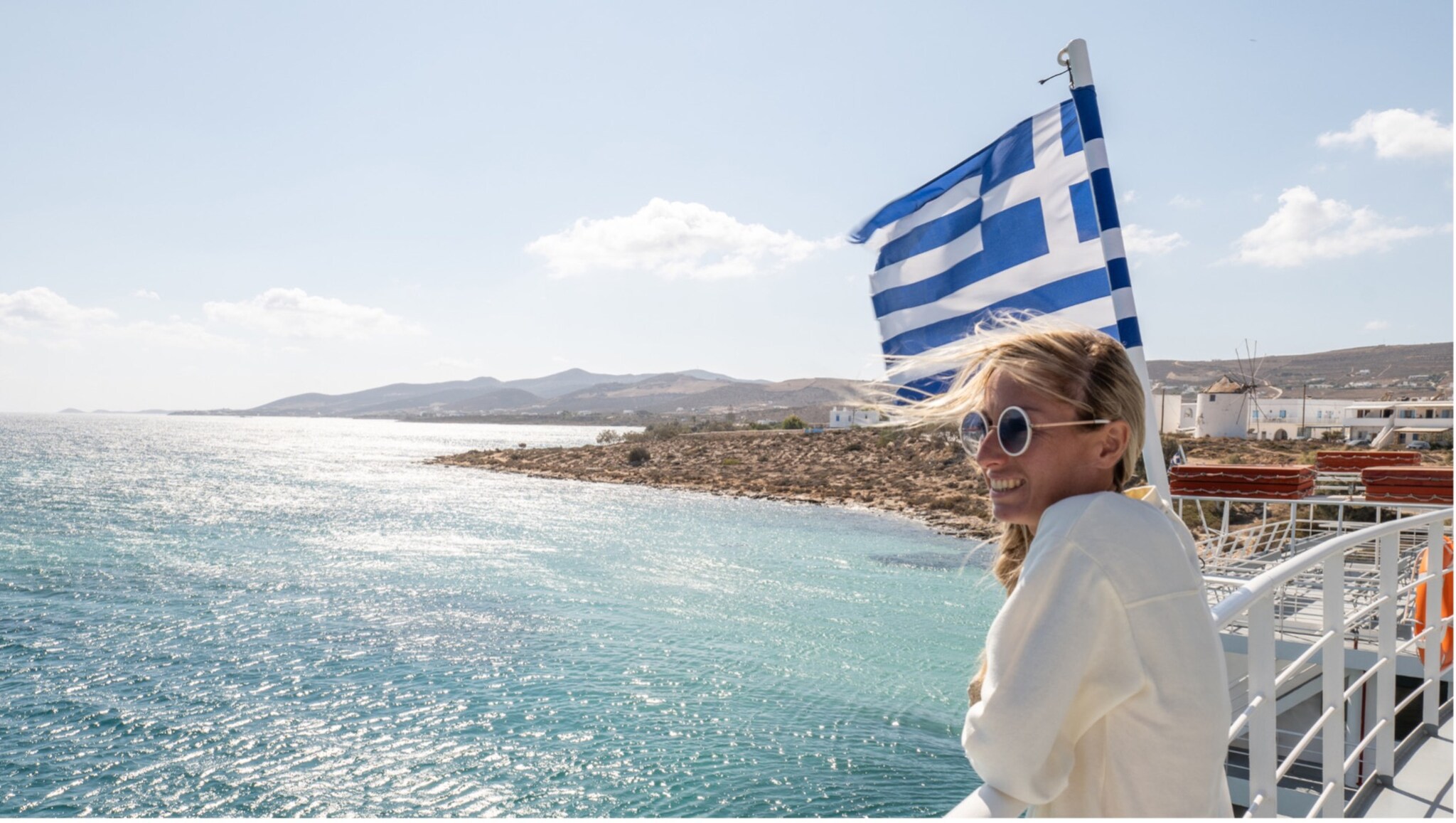 Person lehnt am Geländer einer Fähre mit wehender griechischer Flagge, Blick auf das blaue Meer und eine felsige Küste im Hintergrund