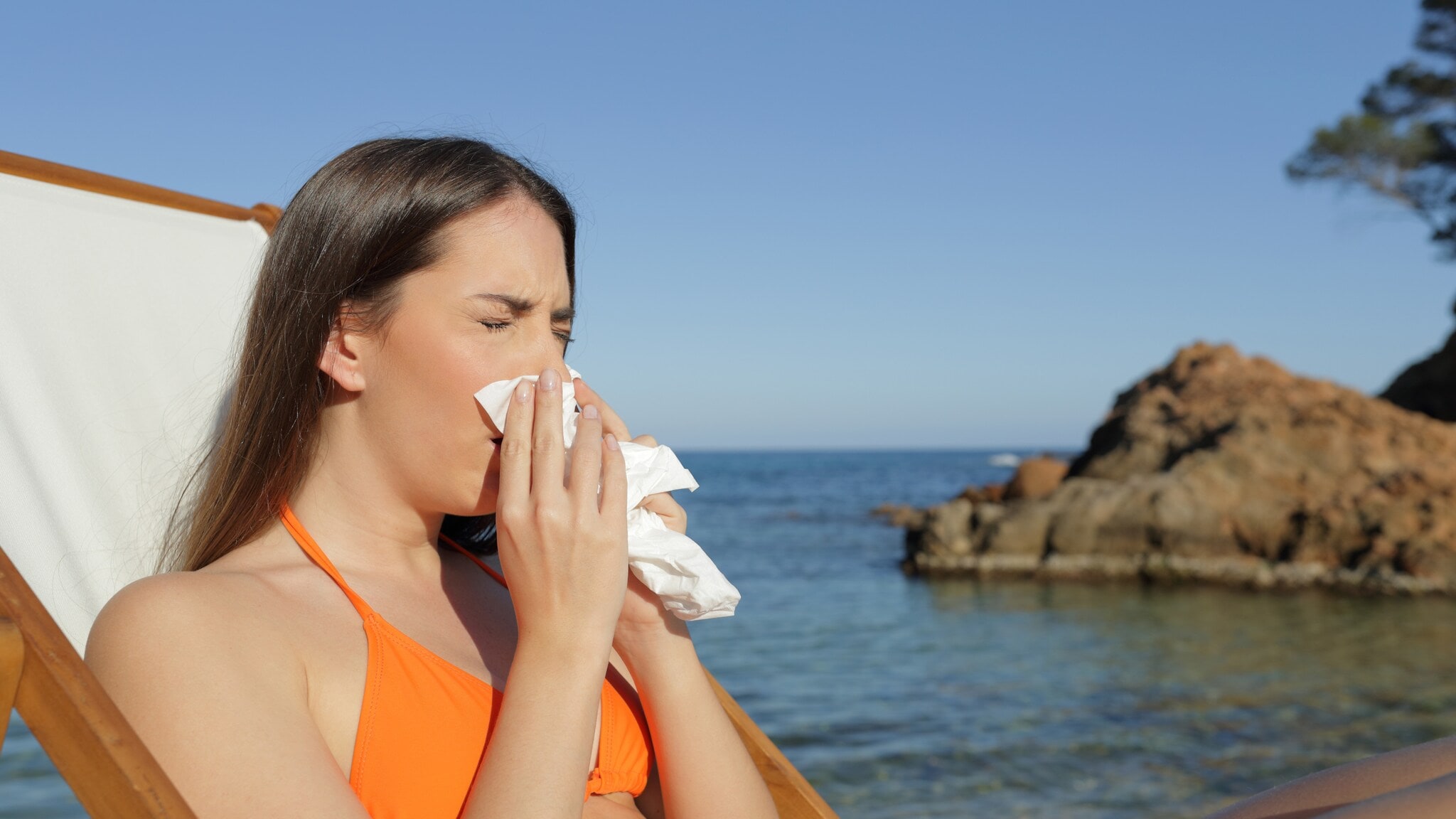 Eine Frau in einem orangefarbenen Bikini sitzt auf einer Liege am Strand und hält ein Papiertaschentuch in der Hand. Im Hintergrund sind Felsen und das Meer sichtbar. Eine Frau in einem orangefarbenen Bikini sitzt auf einer Liege am Strand und hält ein Papiertaschentuch in der Hand. Im Hintergrund sind Felsen und das Meer sichtbar.