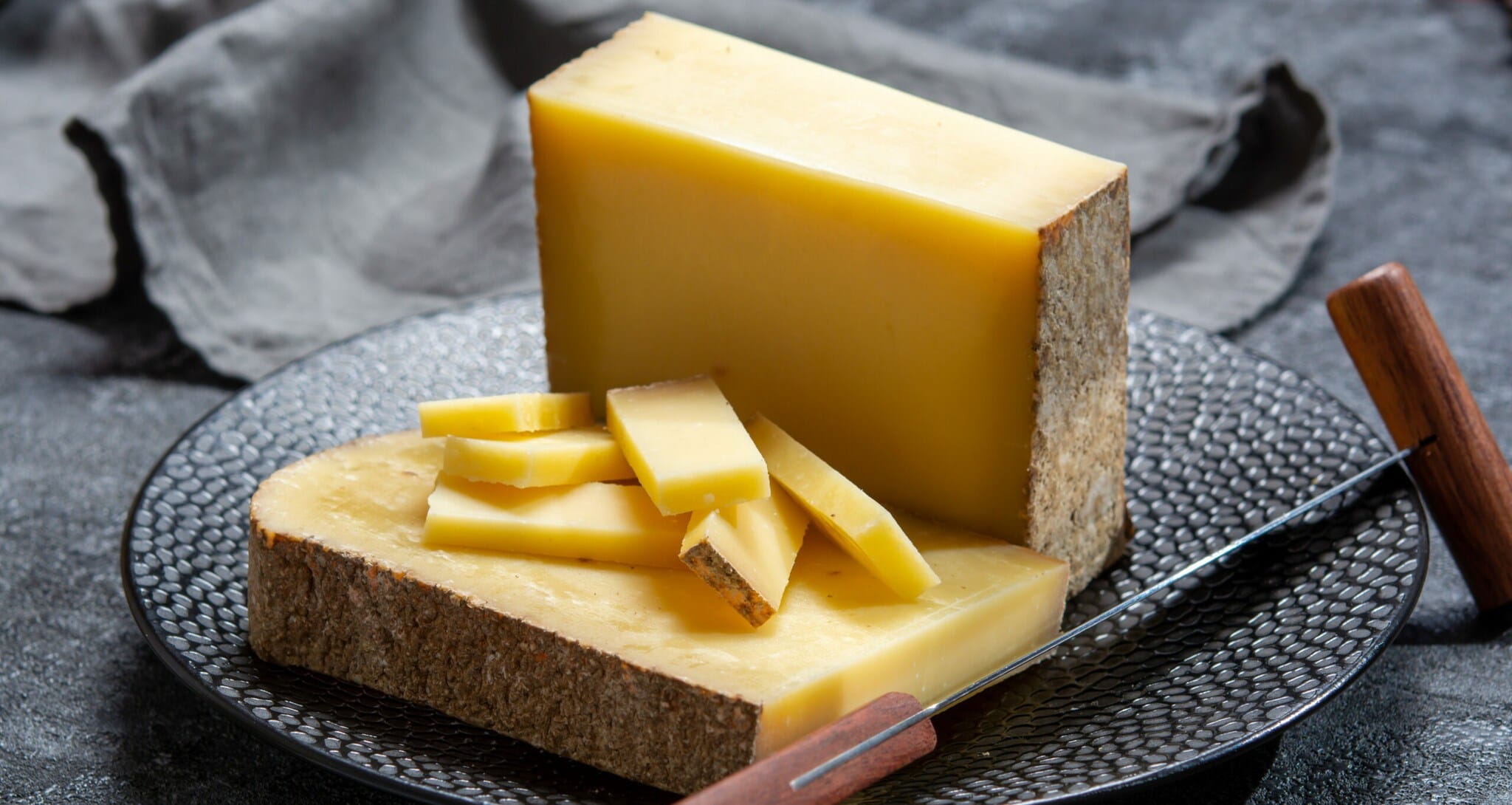 Ein großes Stück Comté-Käse auf einem dunklen Teller, teilweise in Stücke geschnitten, daneben eine Stoffserviette und ein Käsemesser Ein großes Stück Comté-Käse auf einem dunklen Teller, teilweise in Stücke geschnitten, daneben eine Stoffserviette und ein Käsemesser